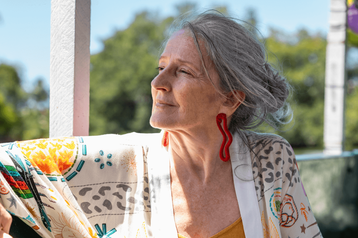 A mature woman with gray hair, dressed in colorful clothing and red earrings, gazing into the distance outdoors with trees in the background.