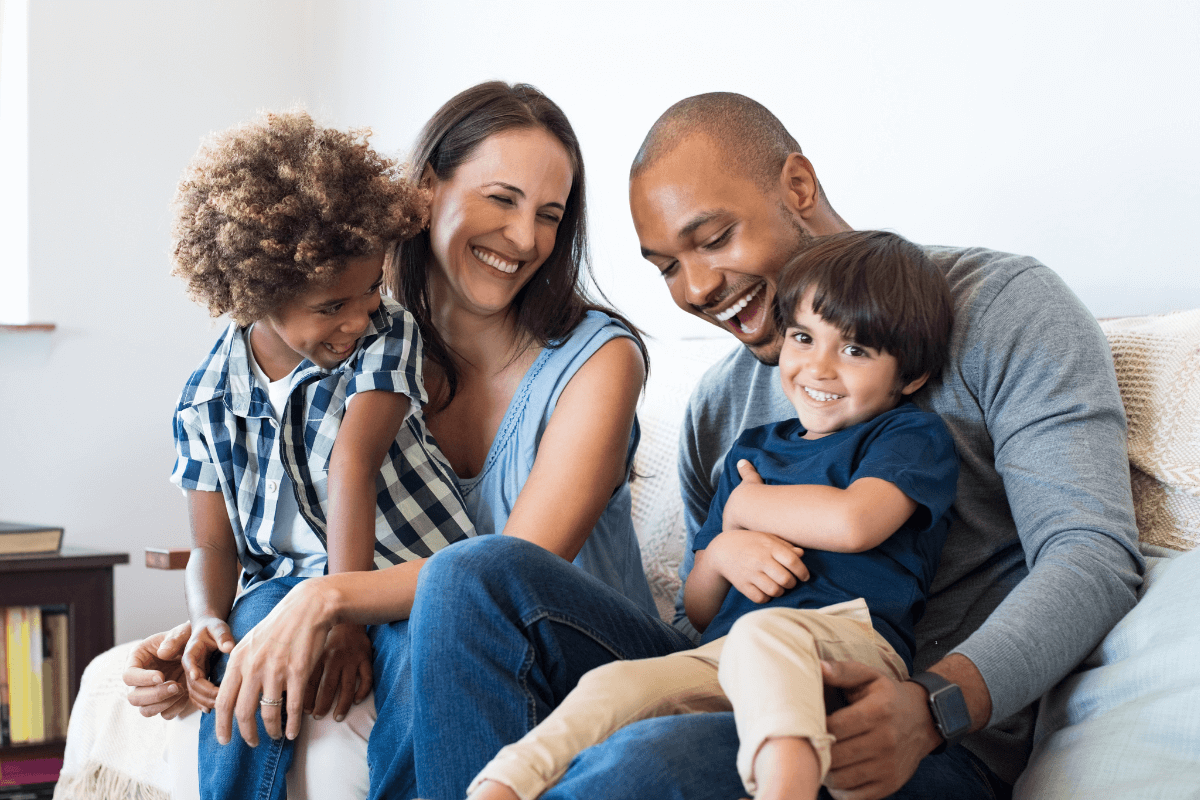 A smiling family of four including a woman, a man, and two young children sitting close together on a couch, enjoying a joyful moment.