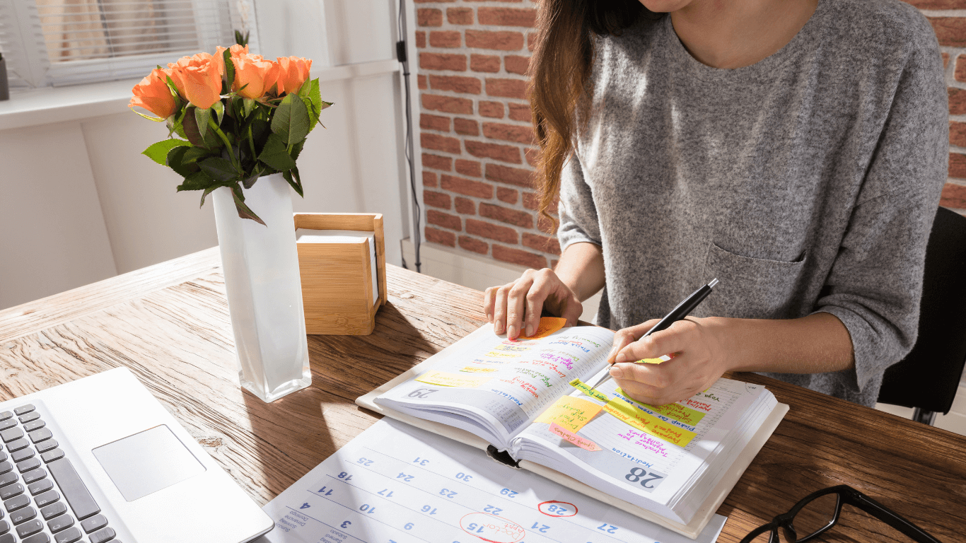 Person writing in a colorful planner on a wooden desk, with a laptop, a vase of peach roses, a pair of glasses, and a calendar visible.