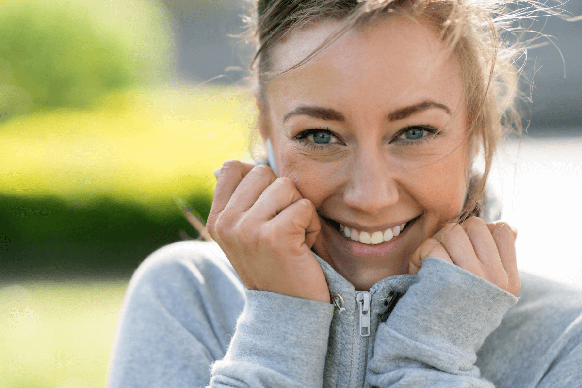 Close-up of a smiling woman with blonde hair and blue eyes, wearing a gray zip-up hoodie, outdoors on a sunny day.