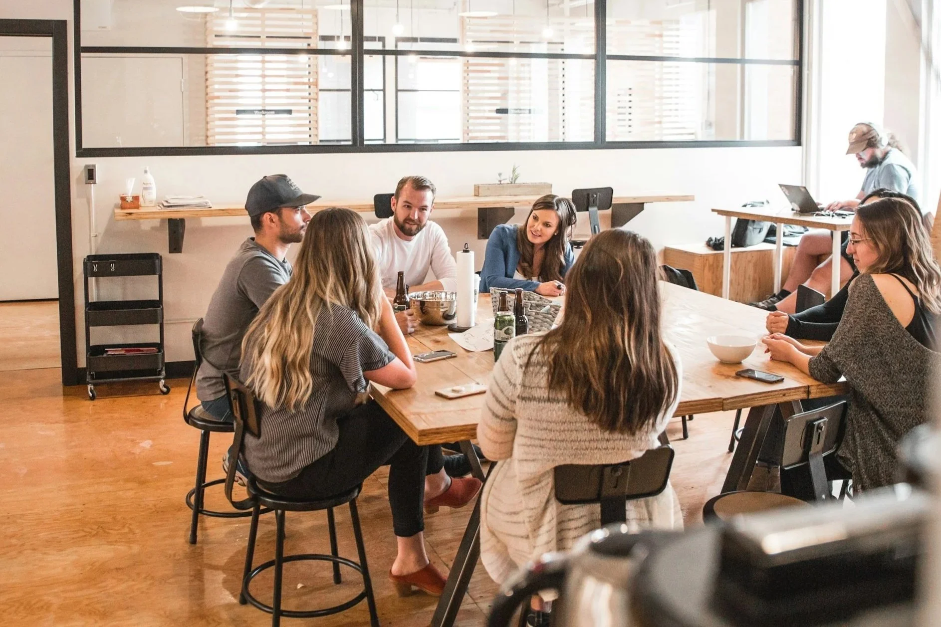 A group of seven young adults sitting around a large wooden table in a modern office or coworking space, engaged in conversation. The room has large windows, wooden floors, and minimal decor, with some working on laptops and drinks on the table.