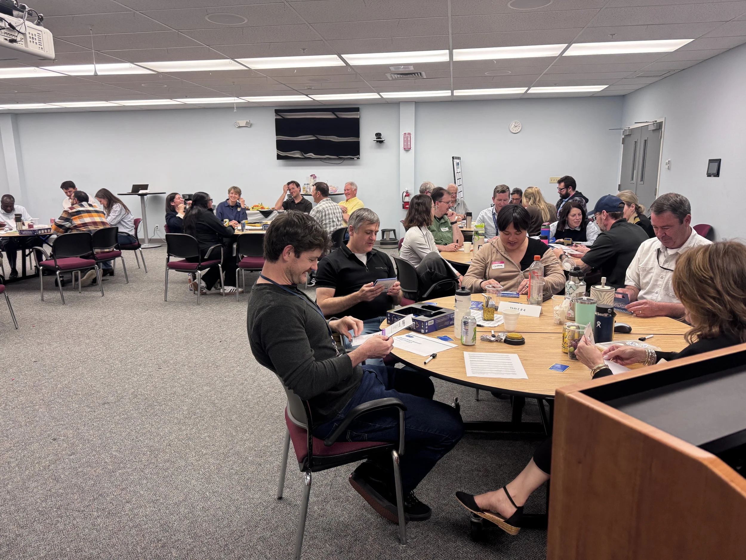 People sitting at round tables in a conference room, some reading, some talking, with snacks and drinks on tables, a large black TV screen on the wall, and a clock showing 2:10.