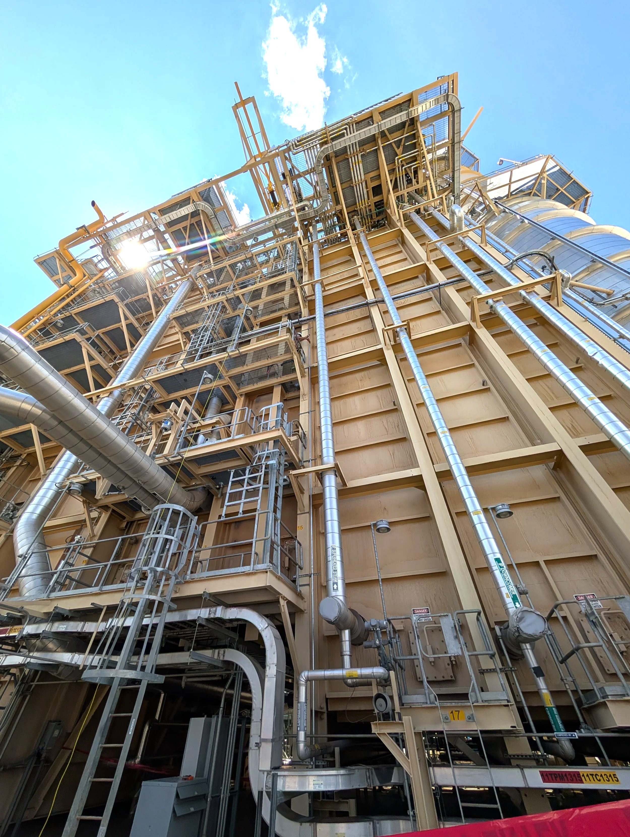 An industrial building with numerous pipes, metal ladders, and support structures against a blue sky with some clouds.