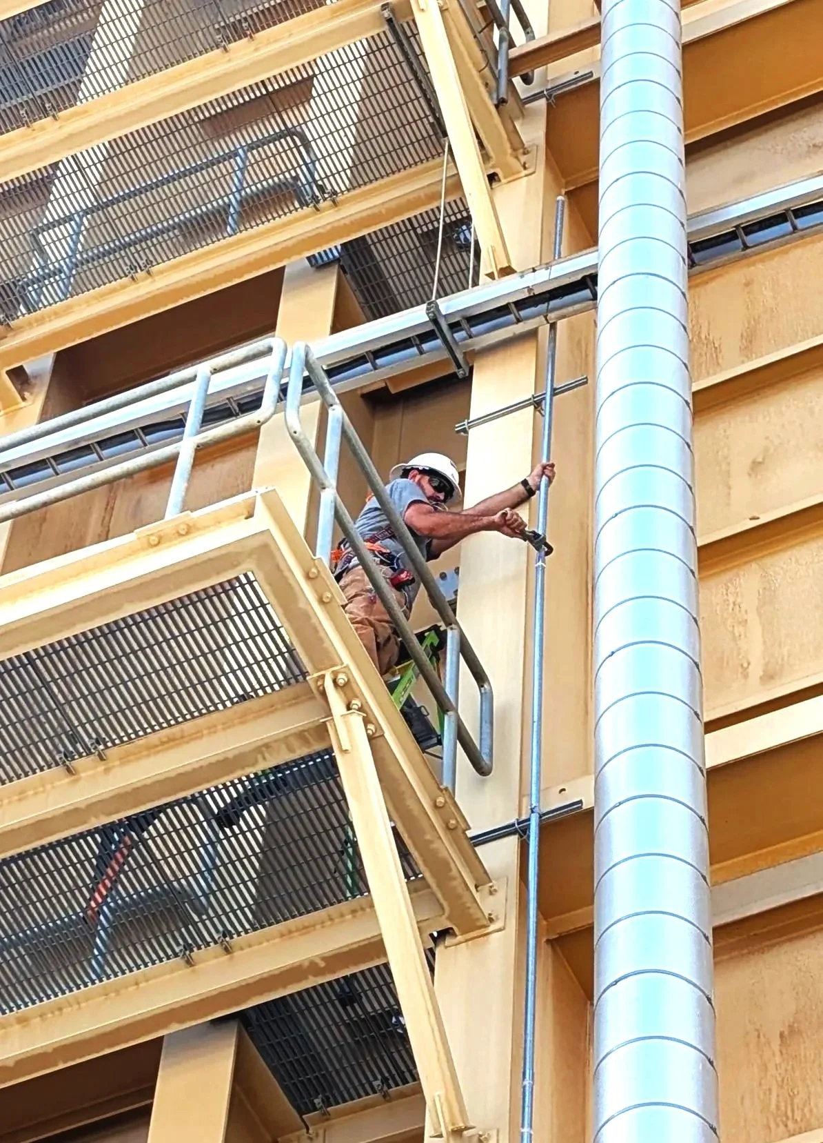 A construction worker wearing a helmet and safety glasses is working on a wooden panel, standing on a scaffold inside a building with exposed wooden and metal framework.