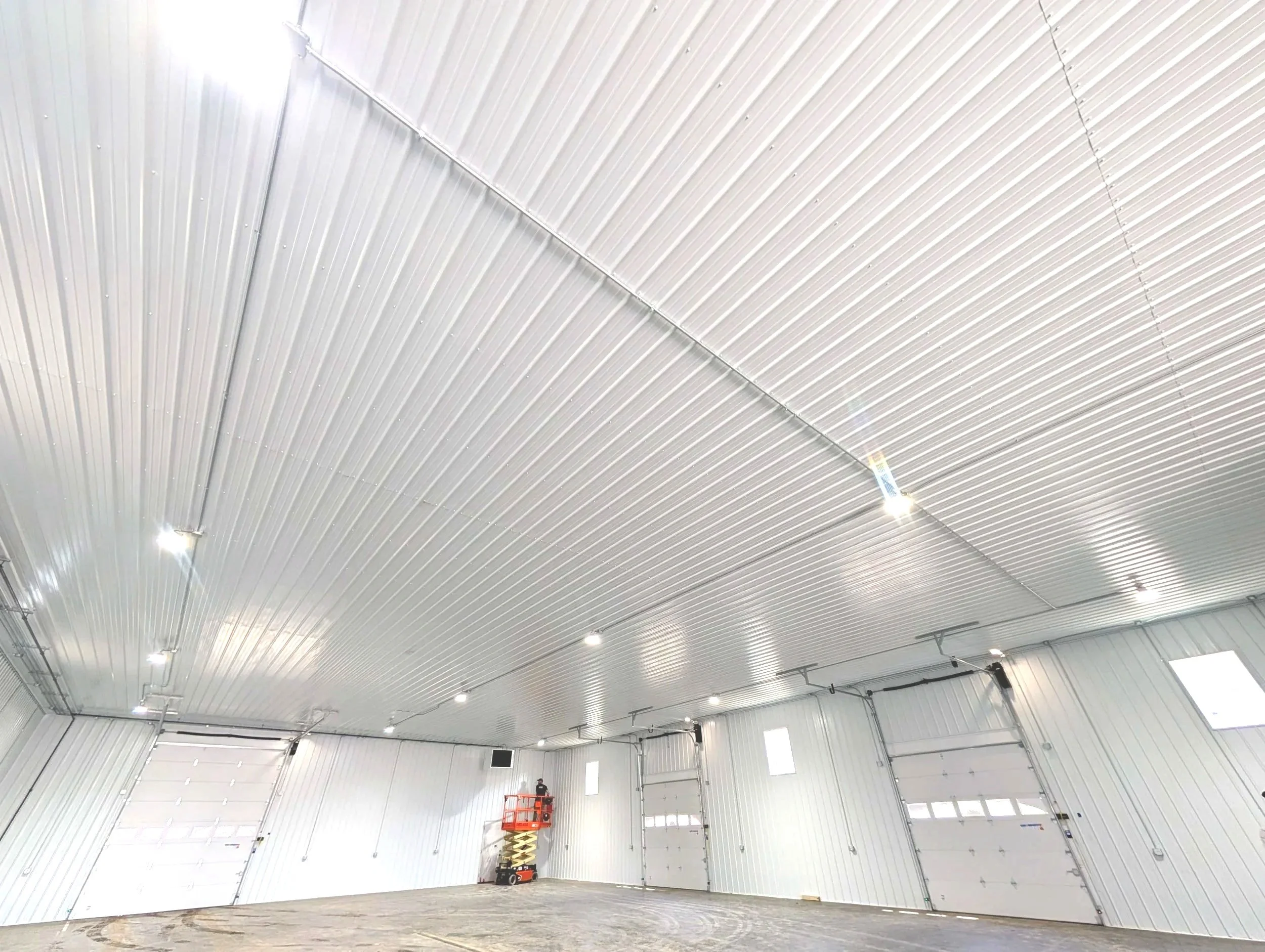 Interior of a large, industrial metal building with high ceiling, white insulated metal panels, bright lighting, a scissor lift, and large garage doors.