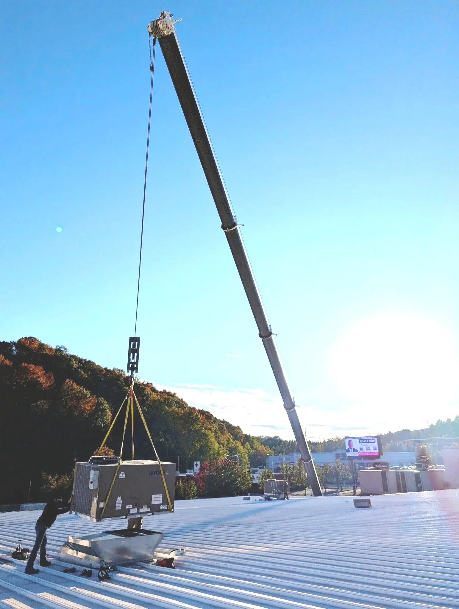 A worker installing or repairing a large rooftop HVAC unit using a crane with a long boom on a metal roof with a forested hill and billboard in the background.