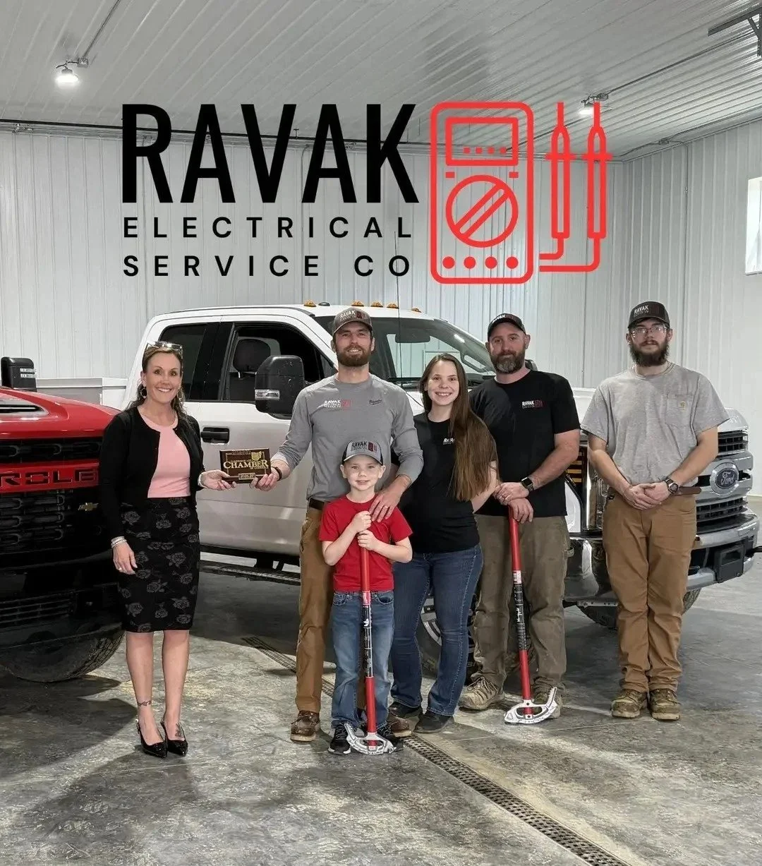 A group of six people standing inside an industrial garage with a pickup truck and another red vehicle. The background features a large sign that reads "RAVAK Electrical Service Co" along with an outline of electrical tools and a multimeter in red. Some people are holding tools, and one person is holding a plaque. Everyone is smiling and wearing casual or work attire.