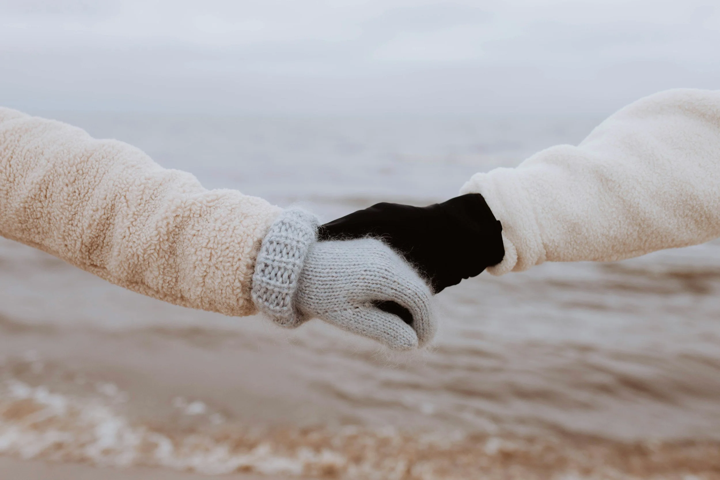 Two people wearing winter gloves holding hands near the shoreline on a cold day.
