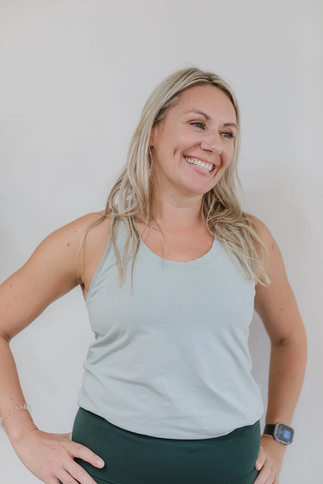 A woman with blonde hair smiling, wearing a light gray athletic tank top and dark green leggings, standing with her hands on her hips against a plain white wall.
