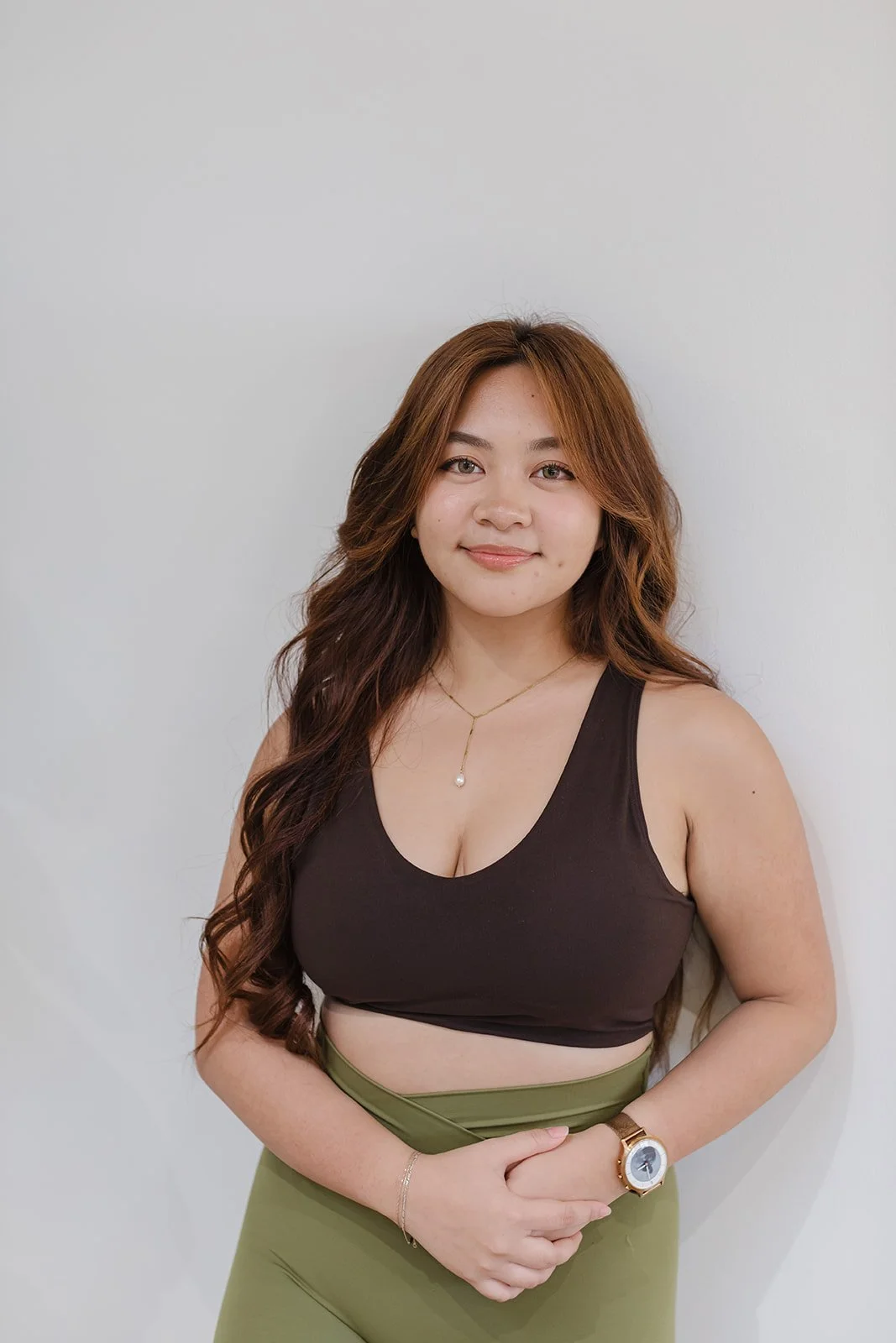 A woman with long, wavy brown hair standing against a white wall, wearing a black sleeveless top, olive green pants, a gold watch, a delicate necklace with a pendant, and a bracelet.