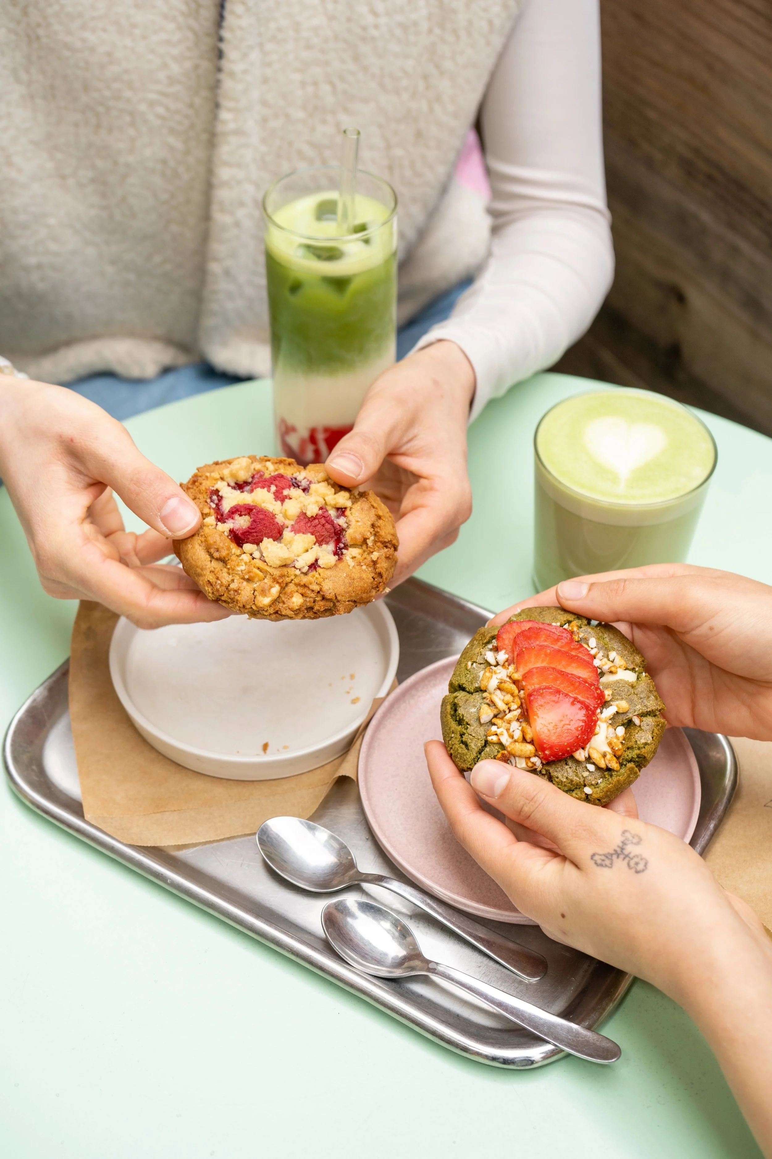 Deux personnes partagent des biscuits, une avec un biscuits aux fruits rouges et l'autre avec un biscuits à la fraise et aux noix, sur un plateau en métal avec deux cuillères, deux boissons vertes avec du lait dans des verres transparents sur une table vert clair.