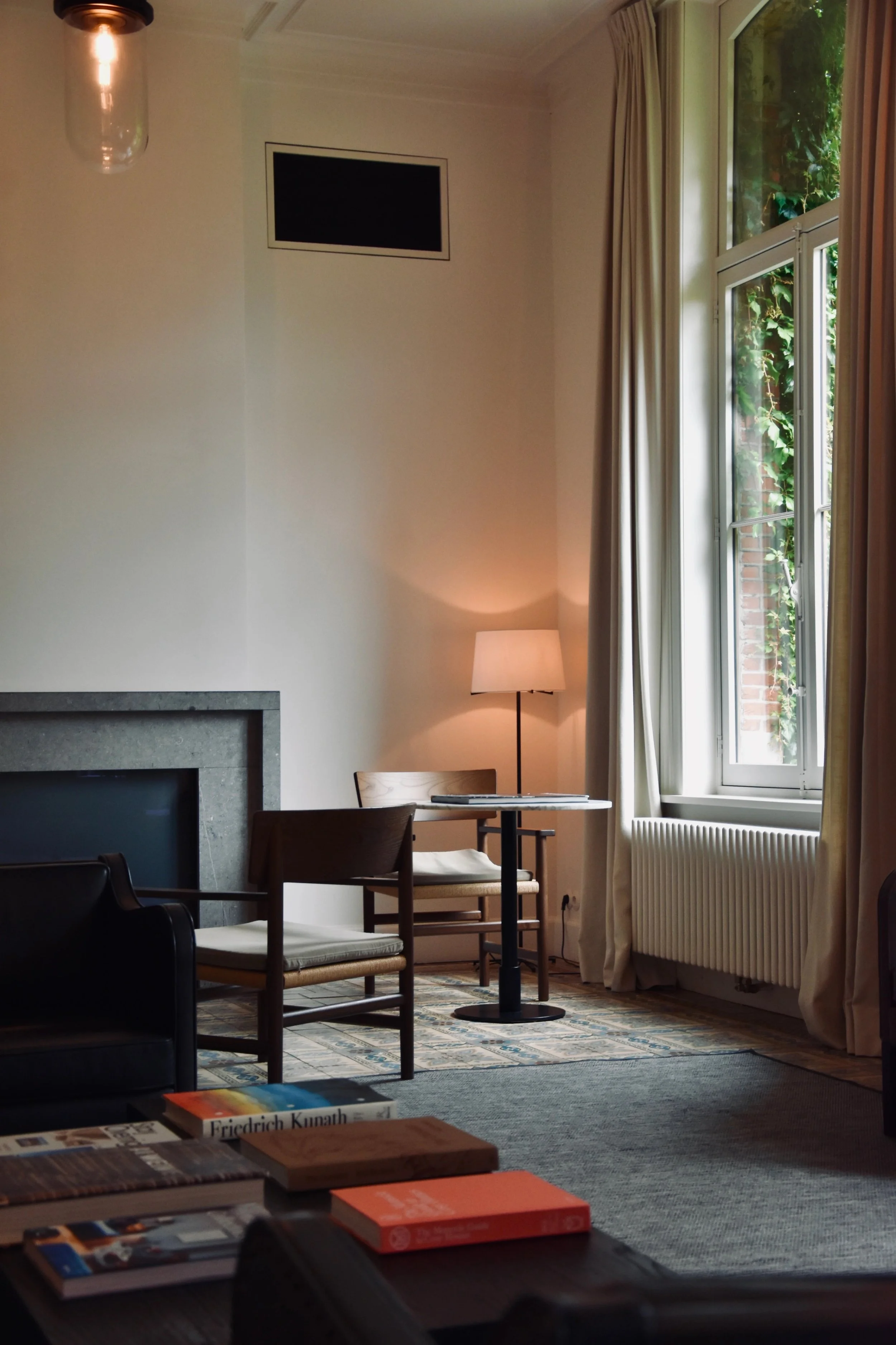 Salon avec un coin lecture, un canapé noir, deux chaises en bois avec coussins, une table ronde, une lampe de chevet allumée, une grande fenêtre laissant entrer de la lumière naturelle, et des livres sur une table