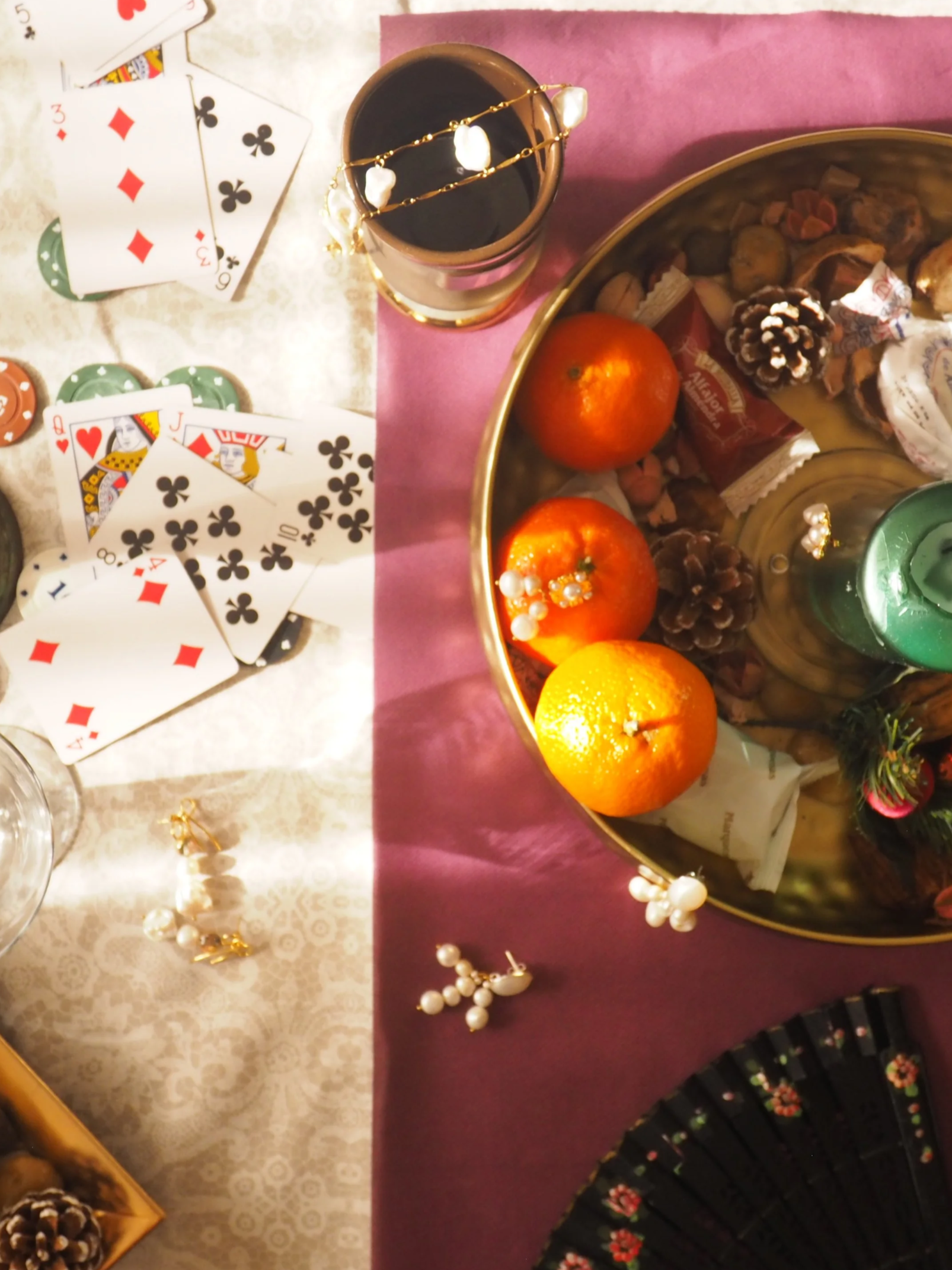 A table decorated for a celebration with playing cards, poker chips, a cup of dark beverage, oranges, and decorative jewelry, including pearl necklaces and a gold chain with pearls.