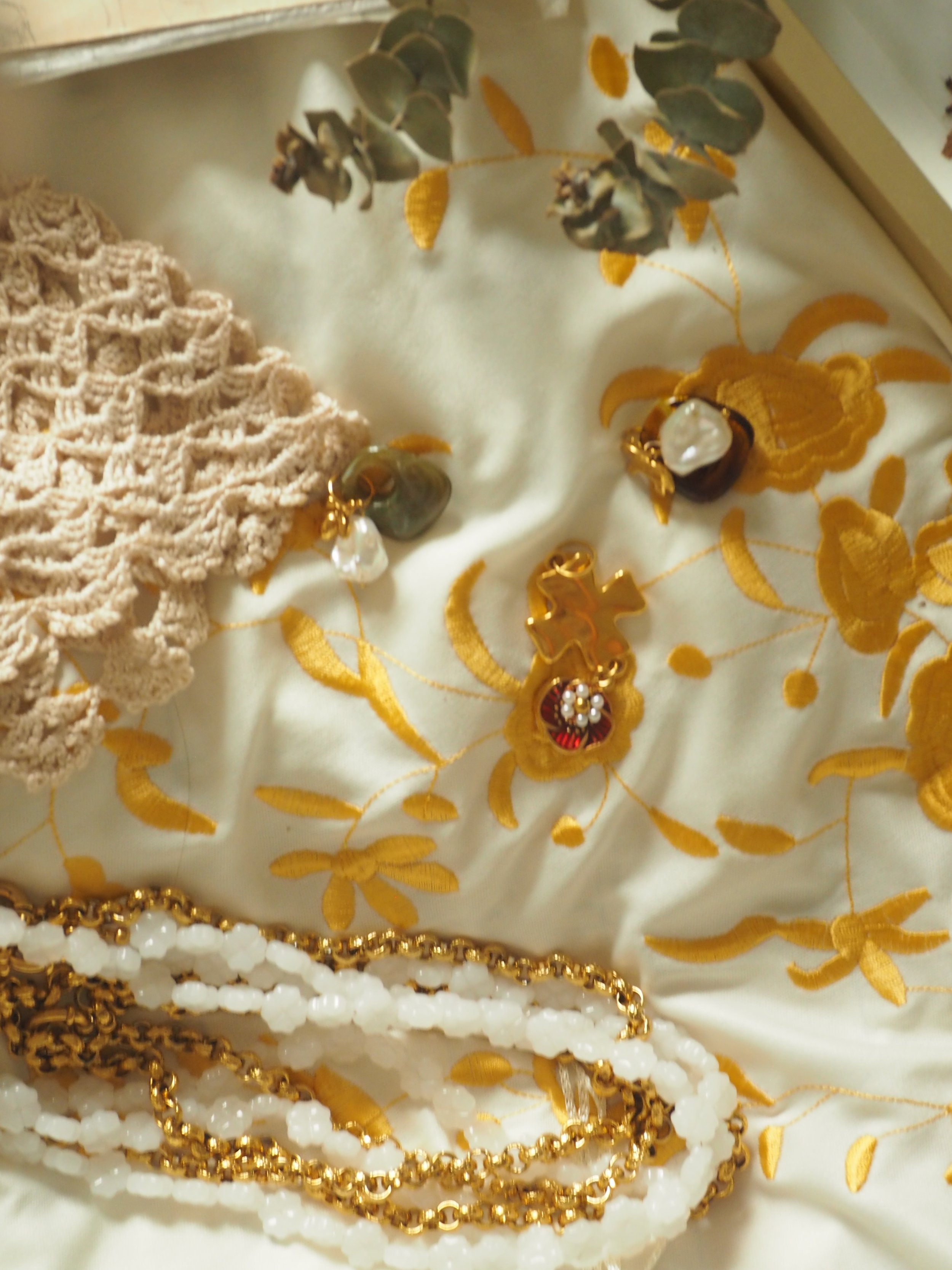Close-up of jewelry, including earrings with pearls and gold chains, on embroidered fabric with yellow floral patterns and lace trim.