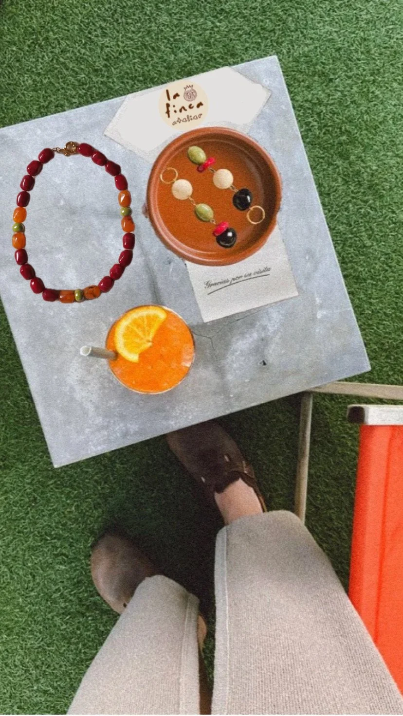 A small grey table with jewelry, a drink with a slice of orange, and a bowl with earrings on it. A person's legs in beige pants and brown shoes are visible.