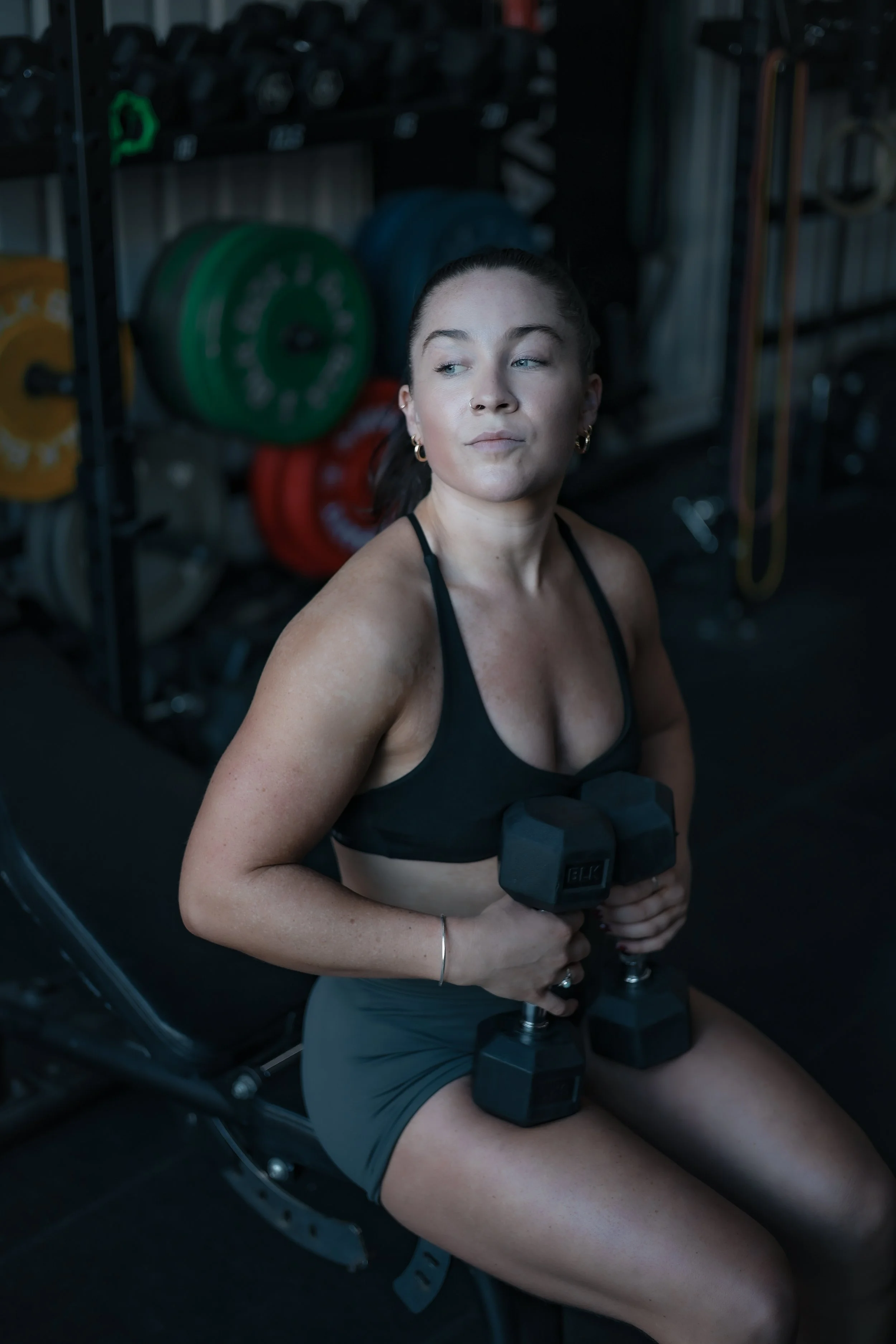A woman in workout clothes, sitting on a workout bench, holding two dumbbells, with weight plates and gym equipment in the background.
