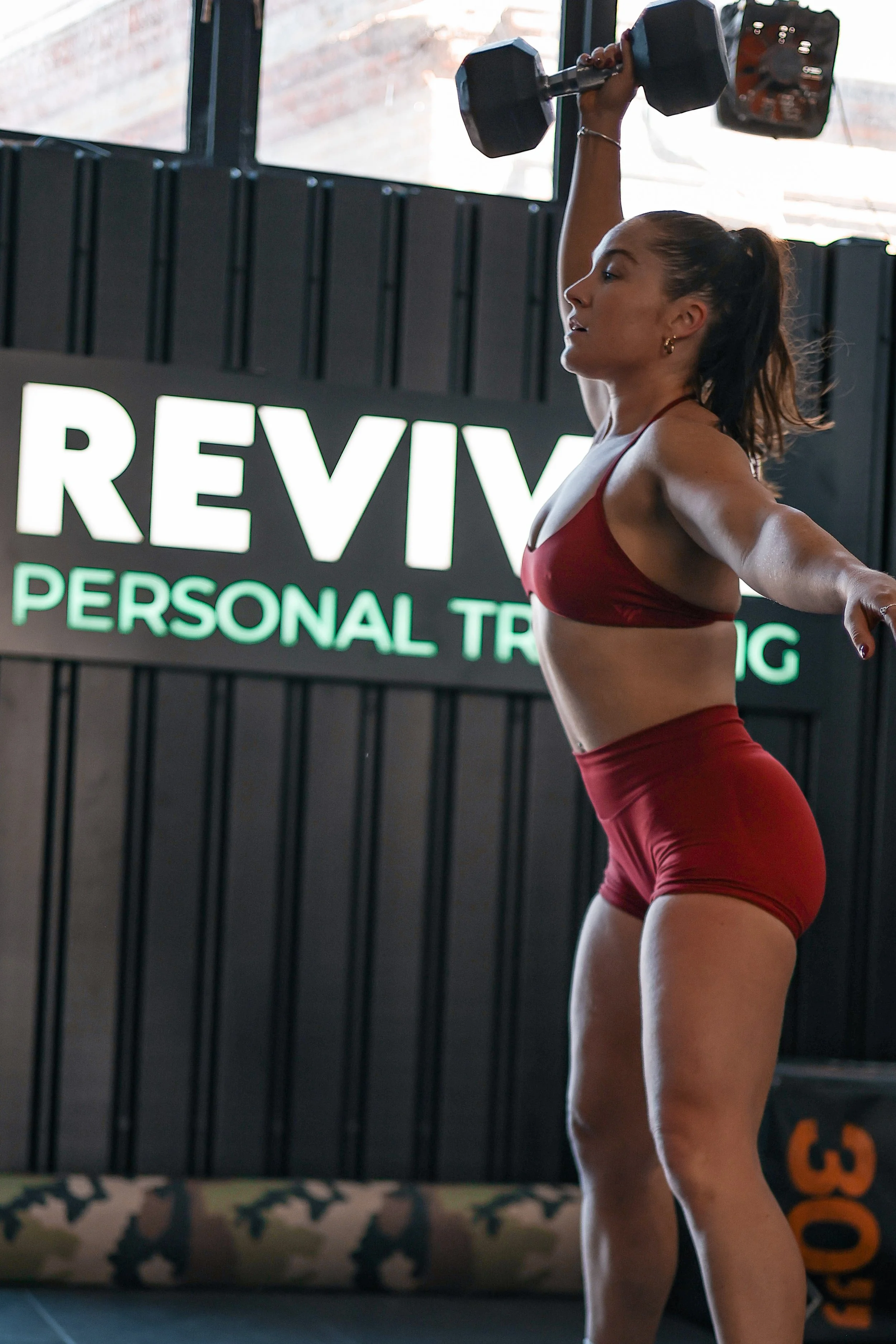 A woman in a red sports bra and shorts lifting a dumbbell with her right arm in a gym with a sign that says "REVI" and "PERSONAL TR."