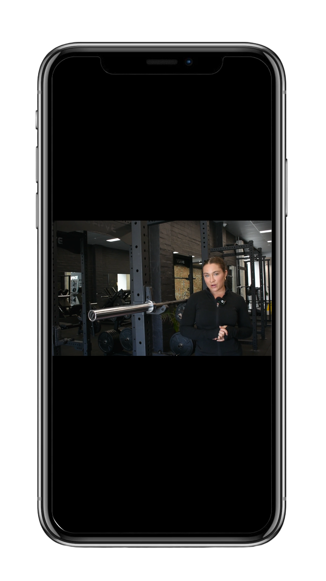 A woman standing in a gym, talking to someone, with gym equipment including a barbell and weight plates around her.