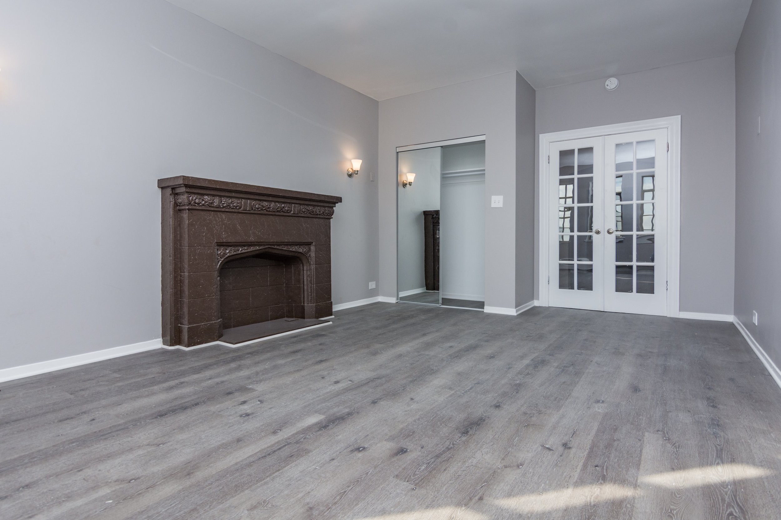 Empty living room with gray walls, a brown fireplace, gray wood flooring, and white double door leading outside.