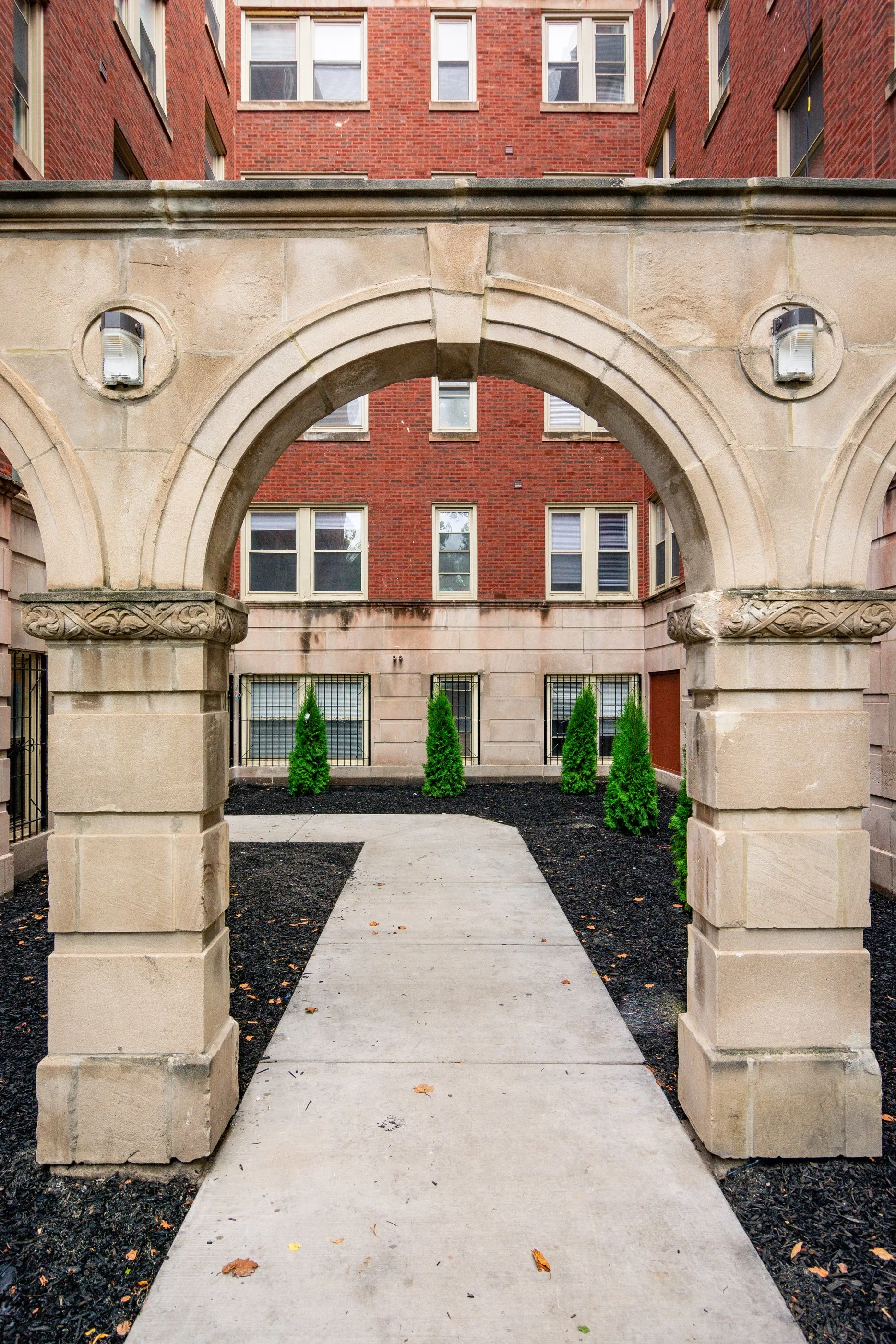 Stone archway leading to a paved pathway with small green trees, surrounded by red brick apartment buildings.
