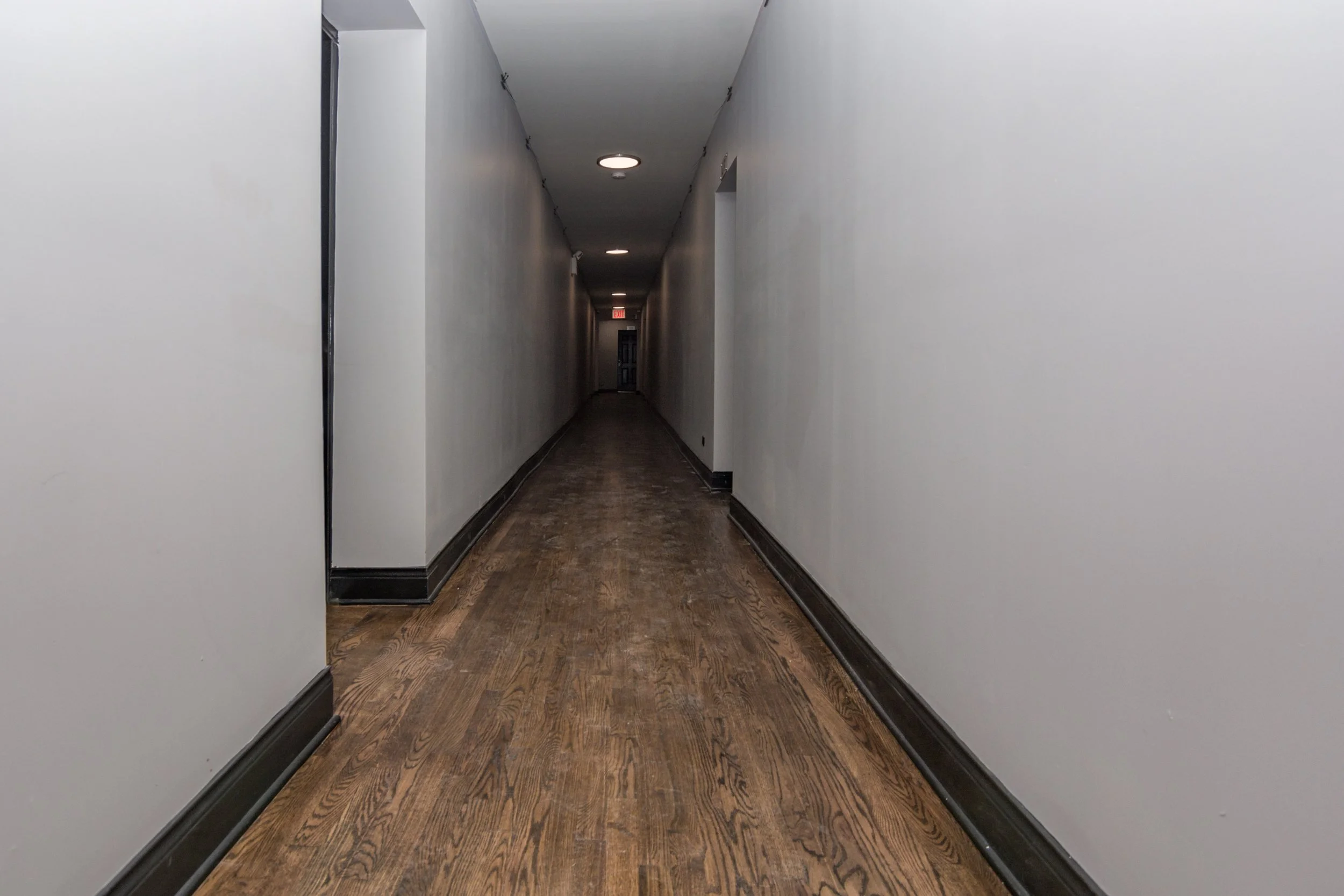 Empty hallway with dark hardwood flooring, white walls, black baseboards, and ceiling lights leading to a door at the end.