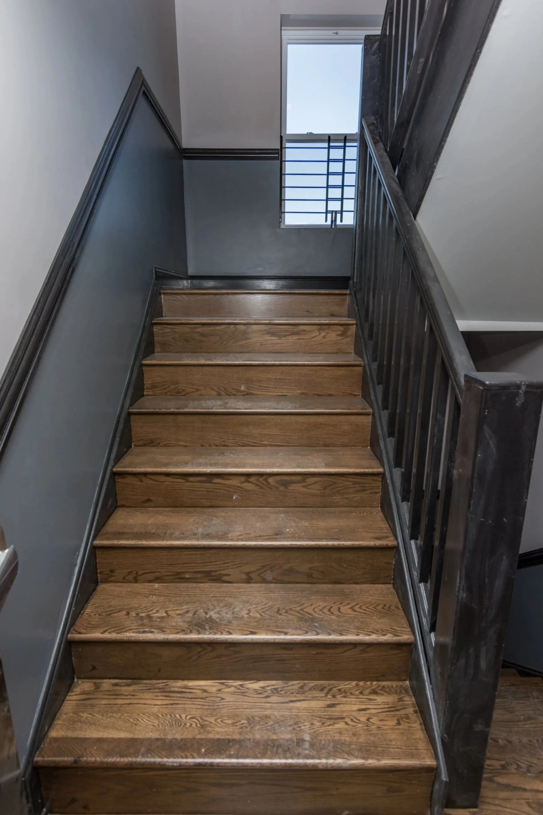Wooden staircase with black railing, gray walls, and a window at the top letting in natural light.