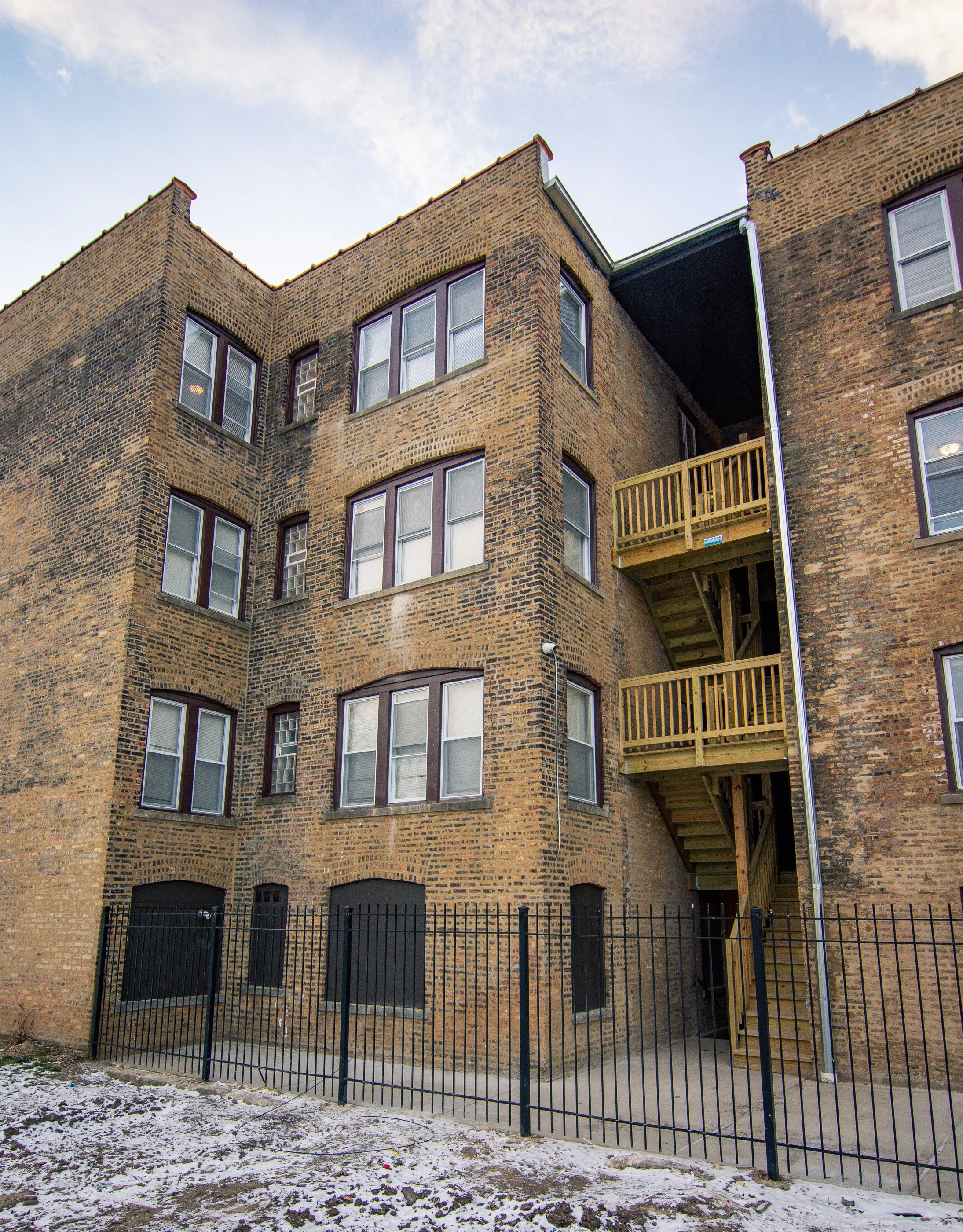 Backside of a brick apartment building with fire escapes and a black metal fence in front, snow on the ground, and a partly cloudy sky.