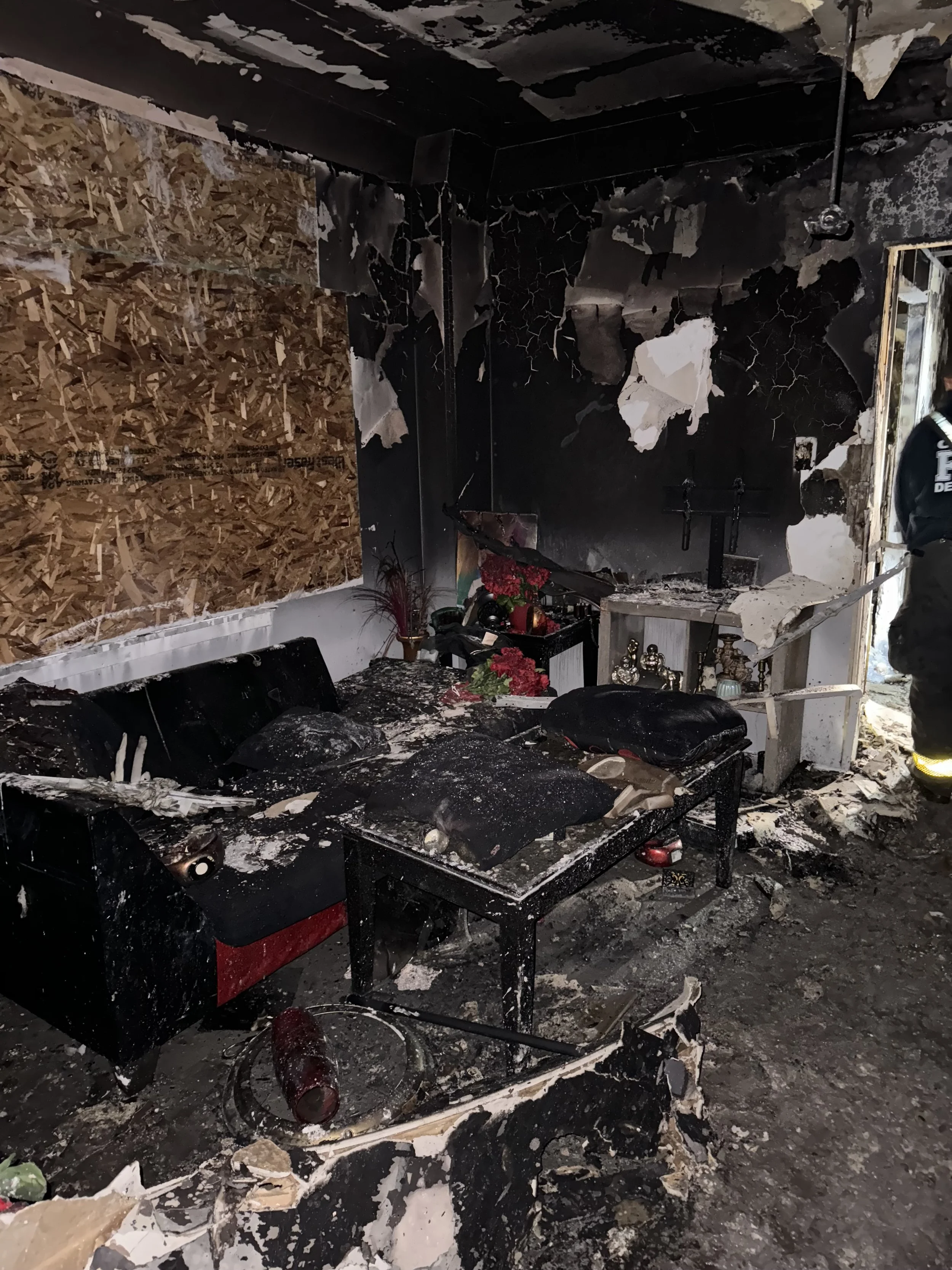 Room with extensive fire damage showing charred walls and ceiling, a burned sofa, a blackened coffee table with debris, and soot covering the floor; interior appears to have been a living space with furniture and decorations also damaged.