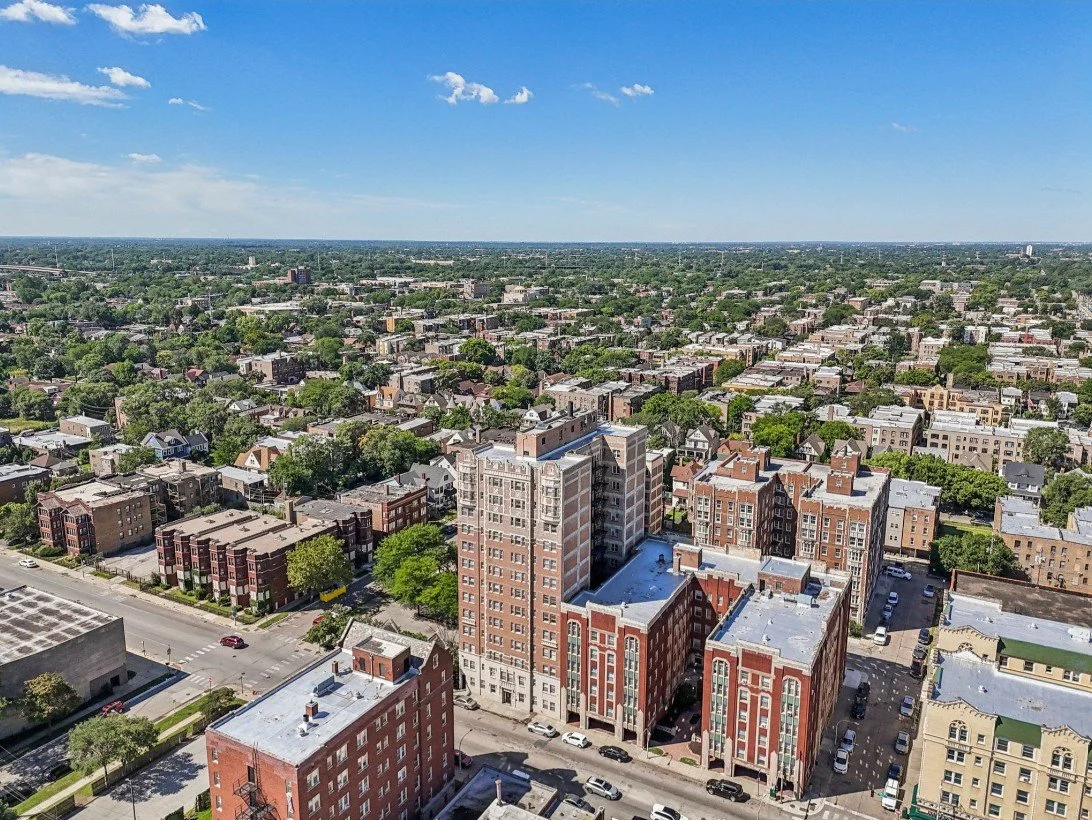 An aerial view of a cityscape with mixed residential and commercial buildings, green trees, and a clear blue sky.