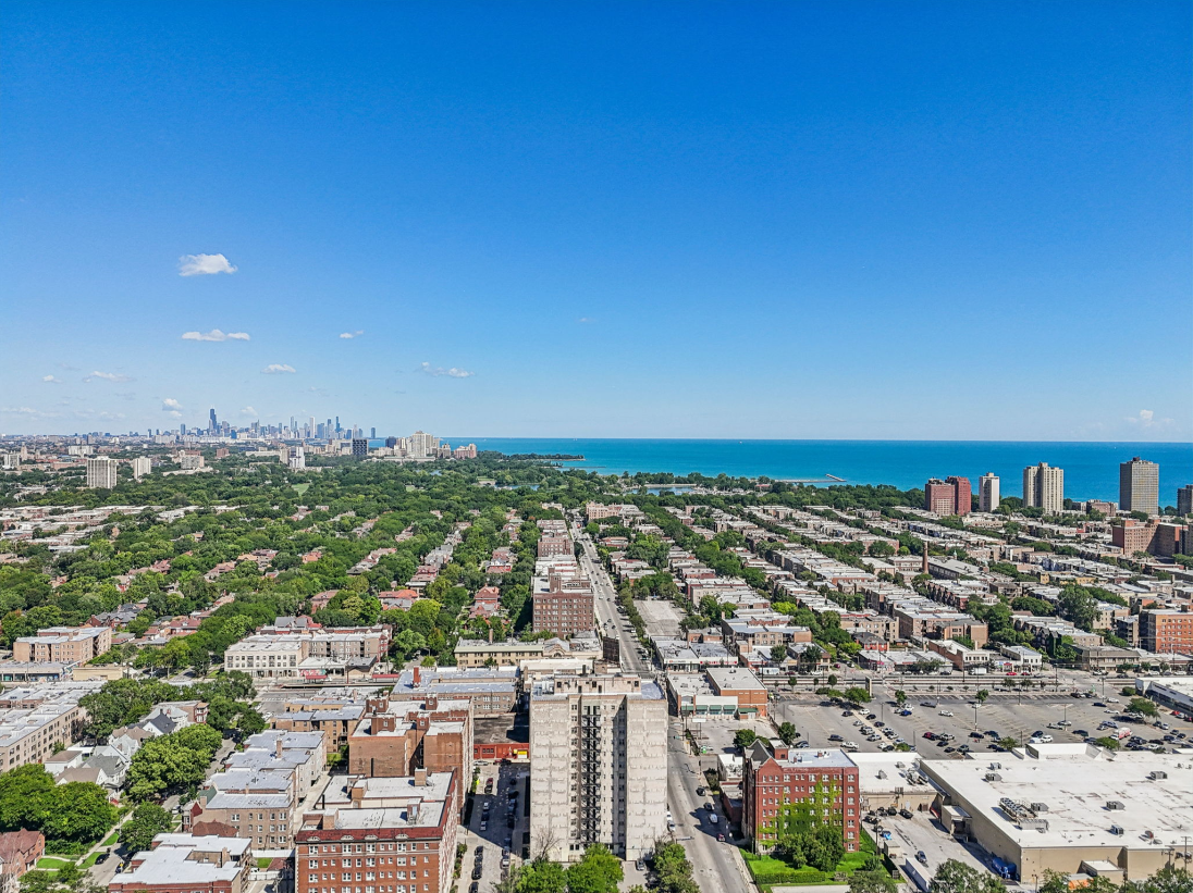 Aerial view of a city with a mix of residential and commercial buildings, green trees, and a large body of water on the horizon on a clear, sunny day.