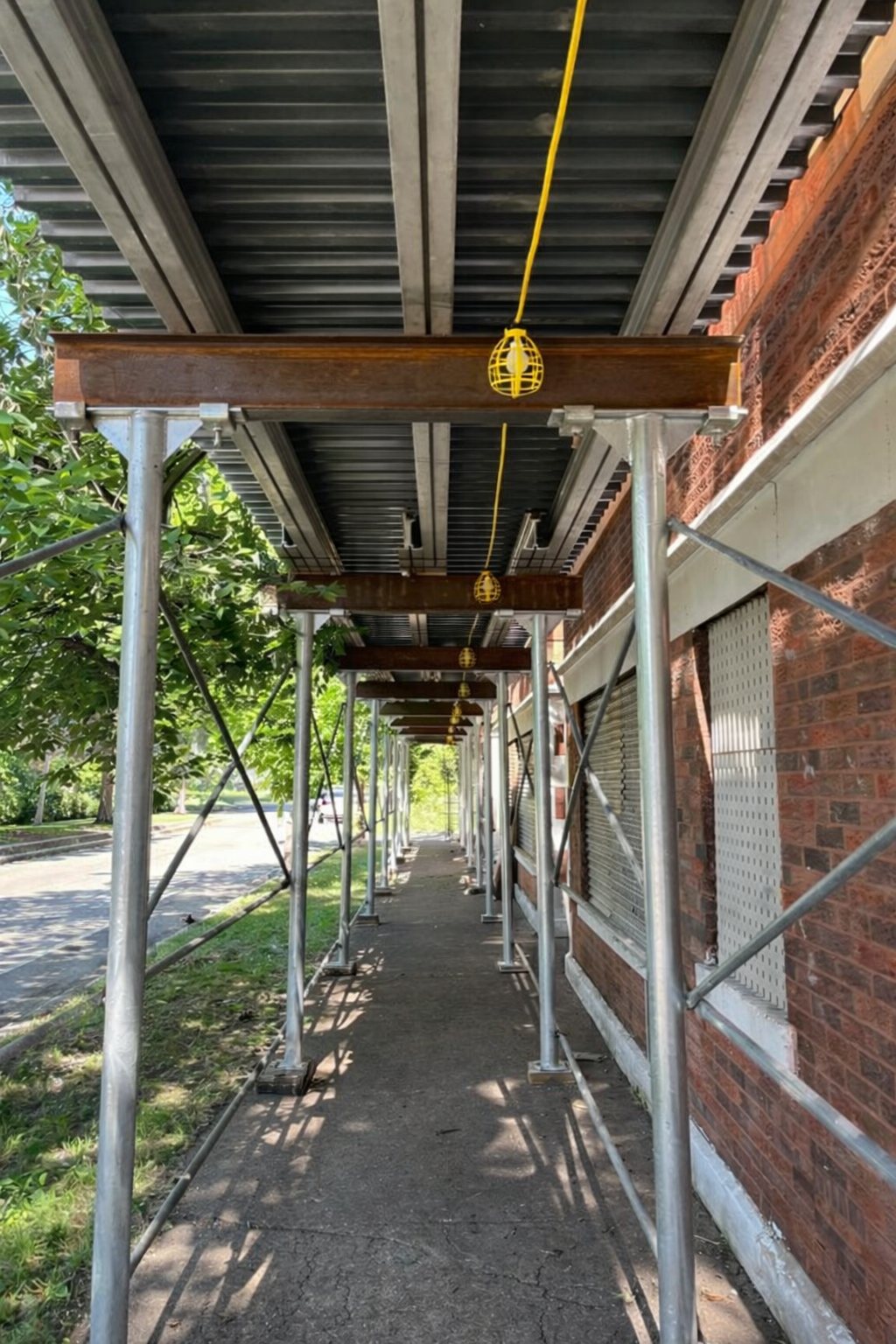 View of a sidewalk alongside a brick building under construction, with scaffolding and yellow construction lights hanging from the ceiling of the scaffolding. Green tree leaves are visible on the left side and the sidewalk stretches into the distance.