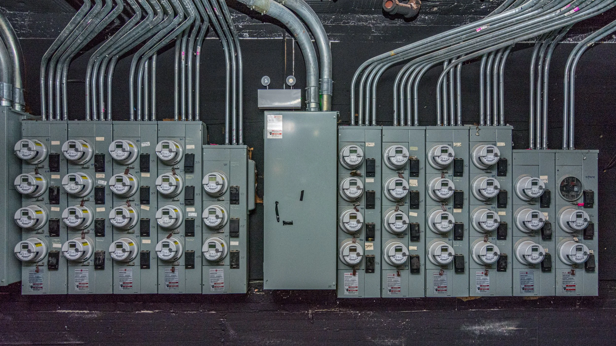 Multiple electric meters and a central electrical box mounted on a wall with numerous conduits running toward the ceiling.