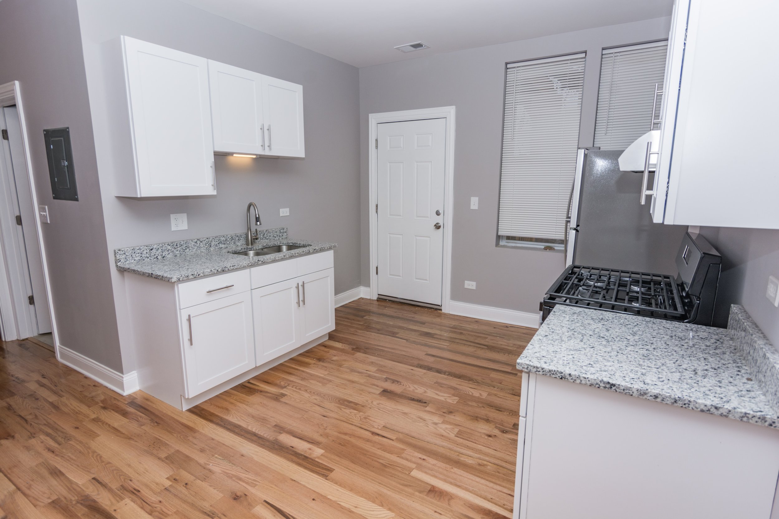 A modern kitchen with white cabinets, granite countertops, a stainless steel sink, a gas stove, hardwood floors, two windows with blinds, a door, and electrical outlets.