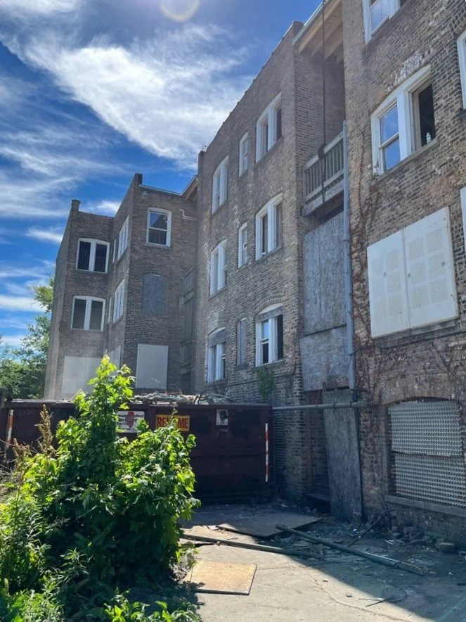 View of a brick apartment building with boarded-up windows and a small balcony, with a blue sky and white clouds above. There is a black dumpster and some debris in the alleyway below.