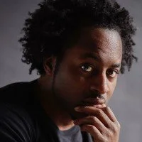Portrait of a man with curly hair and dark skin, looking thoughtfully at the camera with his hand near his mouth.