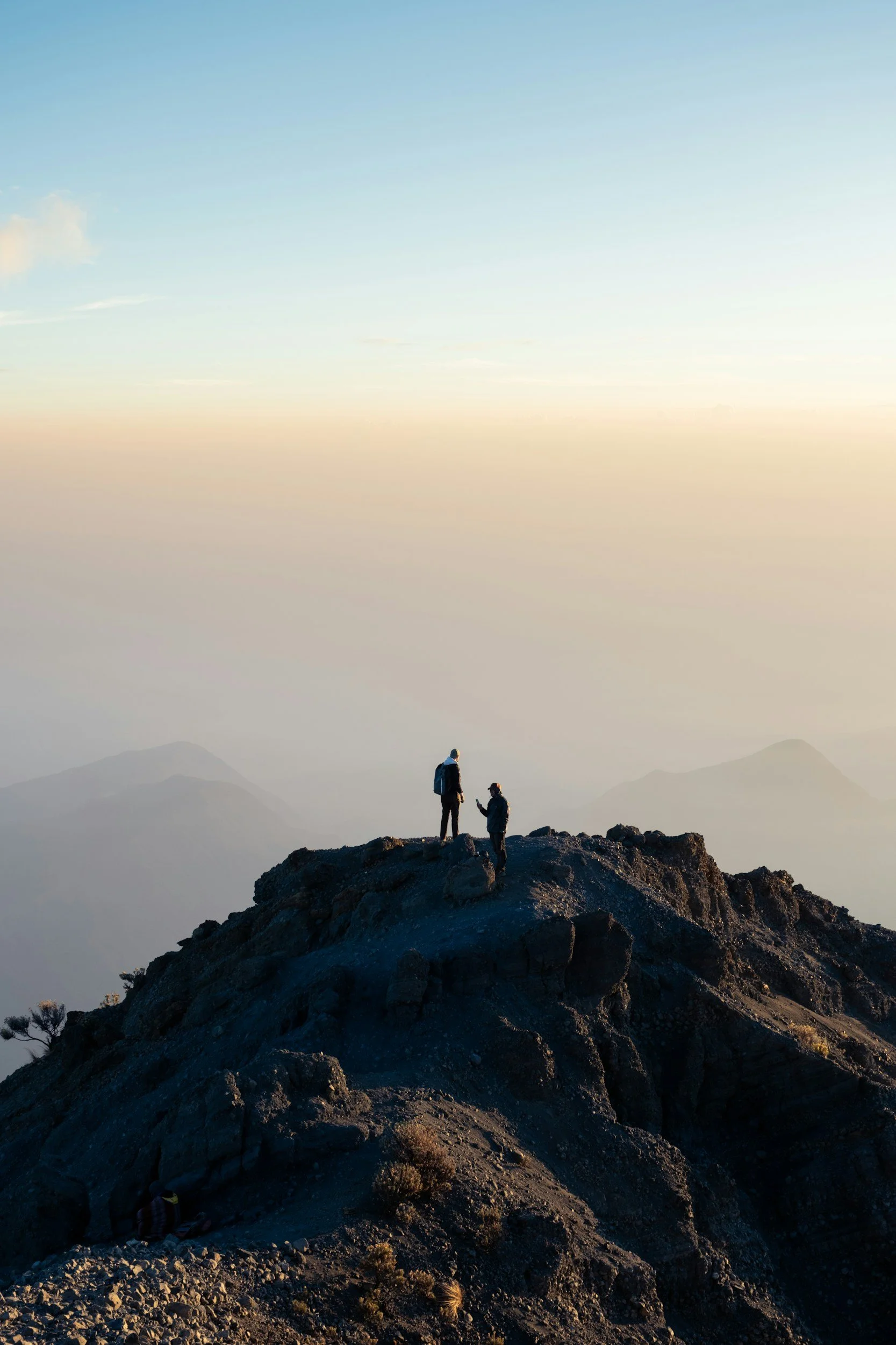 Two hikers standing on a rocky mountain peak during sunrise or sunset, with a misty mountain range in the background.