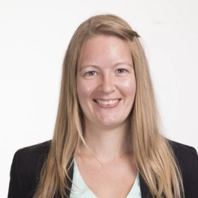A woman with long blonde hair, wearing a black blazer and a light-colored top, smiling against a plain white background.