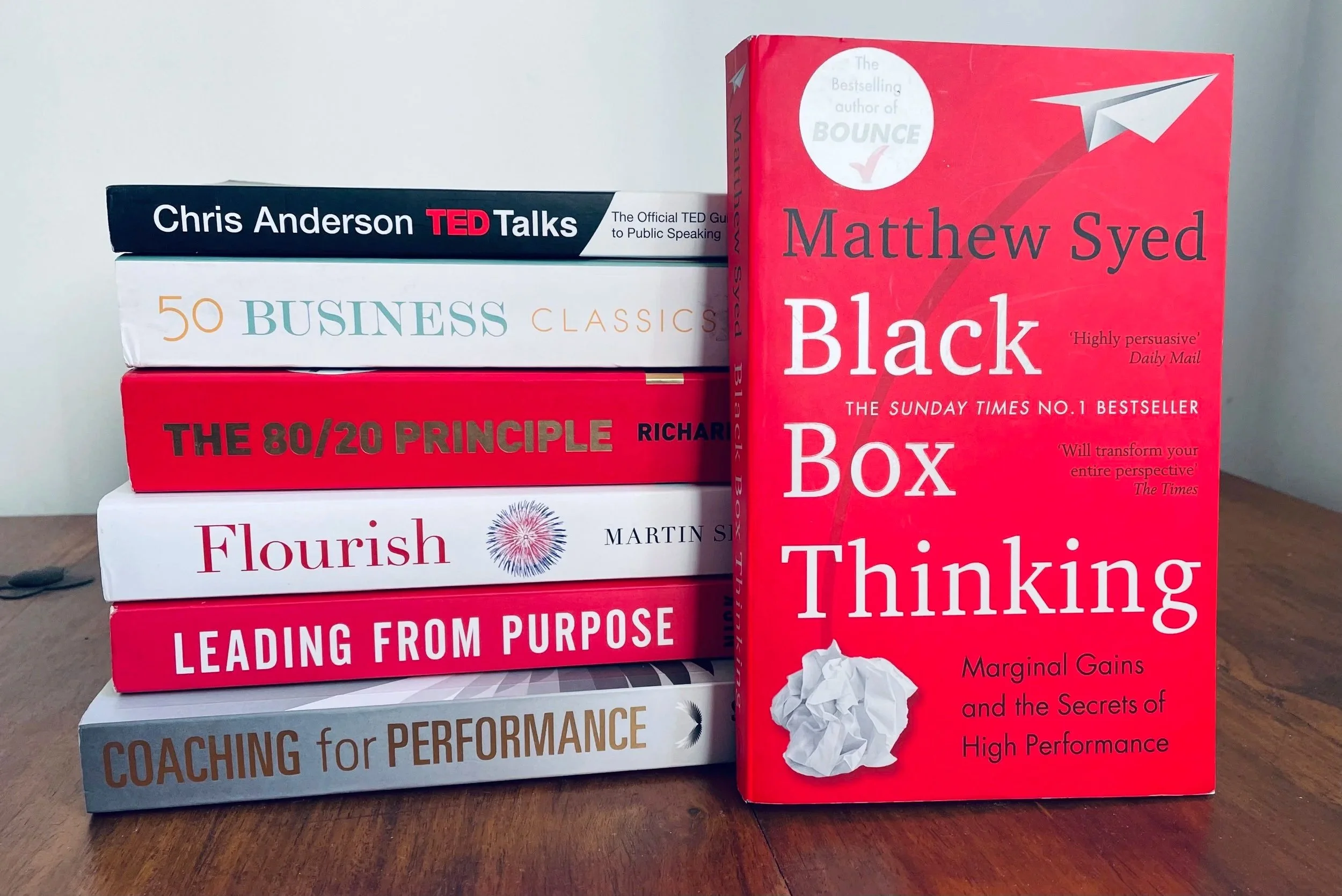 Stack of six books with title of the book on the front cover. The visible titles are "TED Talks" by Chris Anderson, "50 Business Classics", "The 80/20 Principle" by Richard Koch, "Flourish" by Martin Seligman, "Leading from Purpose", and "Coaching for Performance". A seventh book titled "Black Box Thinking" by Matthew Syed stands upright beside the stack.