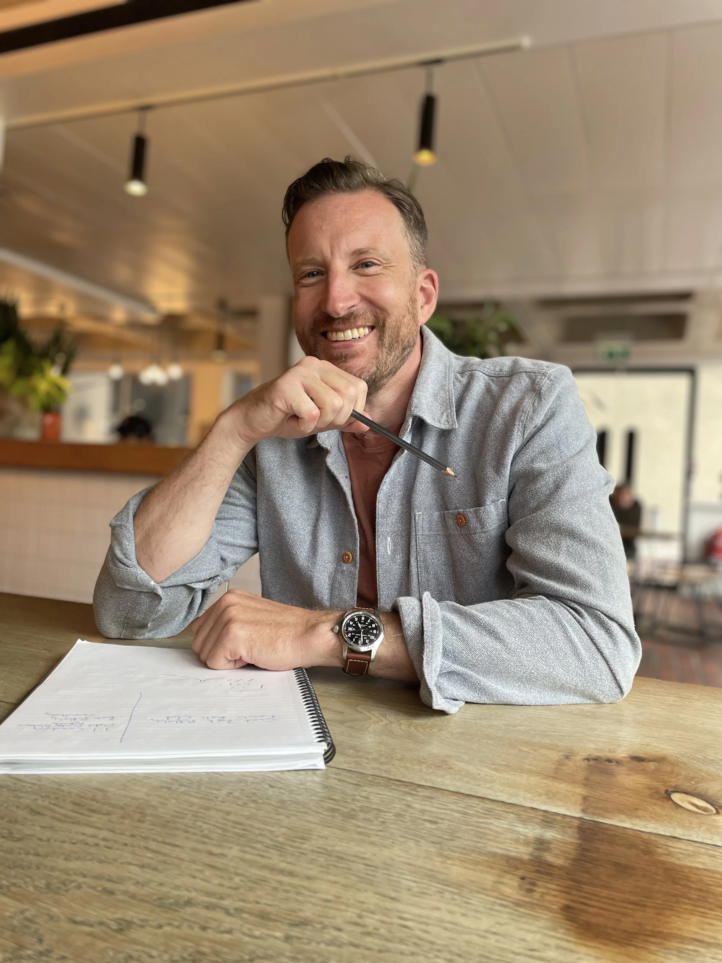 Ben Slight sitting at a wooden table in a cafe, holding a pen near his face, with a notebook open in front of him.