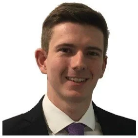 A young man with short dark hair, wearing a dark suit, white shirt, and purple tie, smiling against a plain white background.
