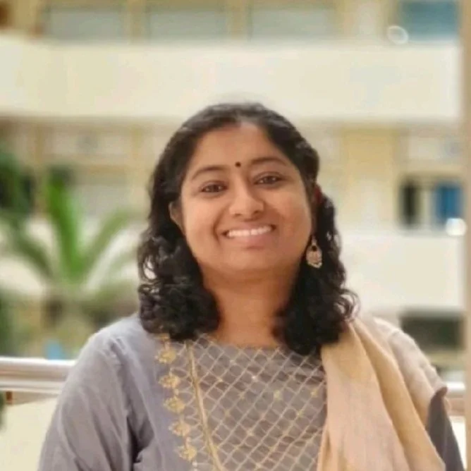A smiling woman with dark curly hair, wearing traditional Indian attire with embroidery, earrings, and a small bindi, outdoors with a blurred background of a building and greenery.