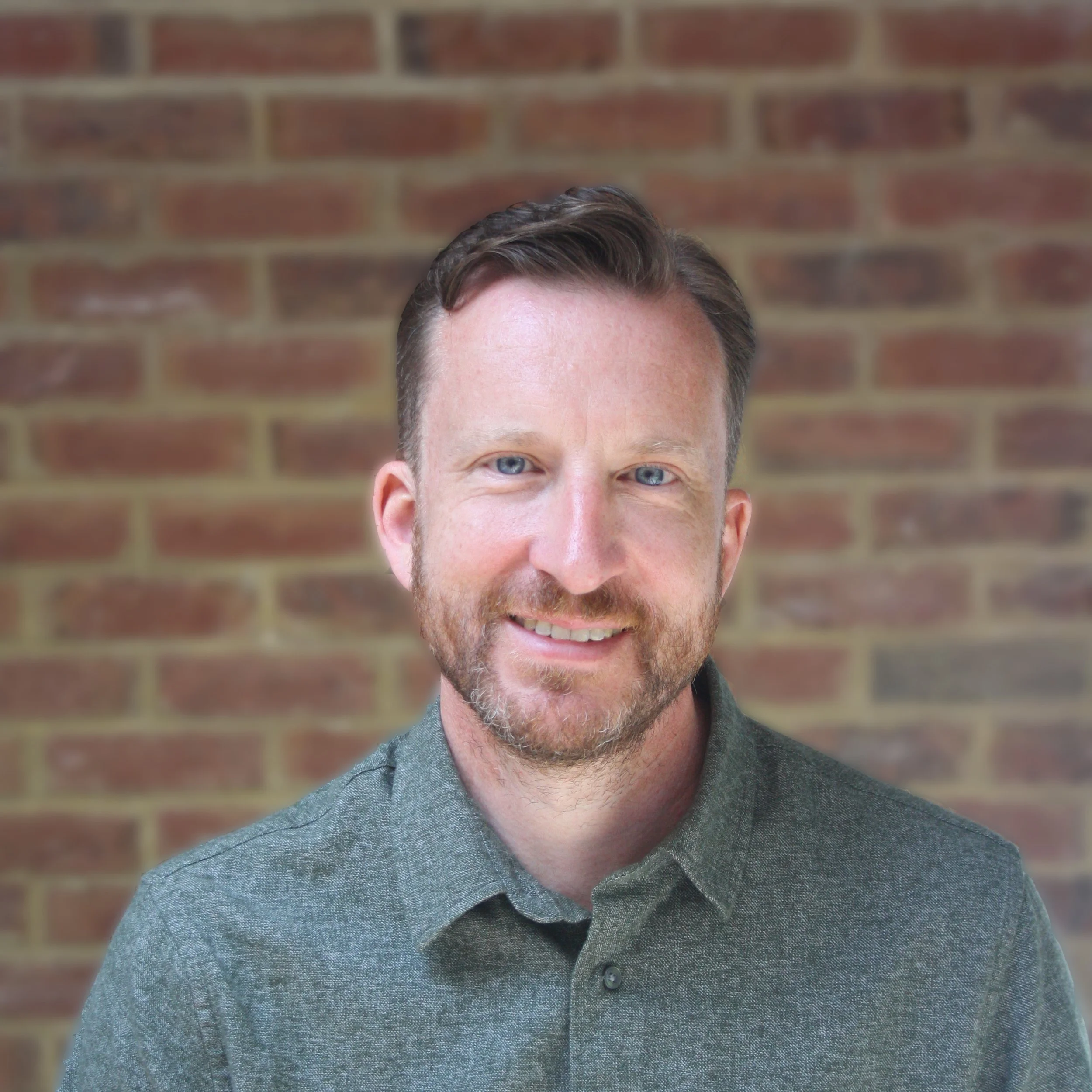 Ben Slight, smiling, in front of a brick wall, wearing a gray button-up shirt.