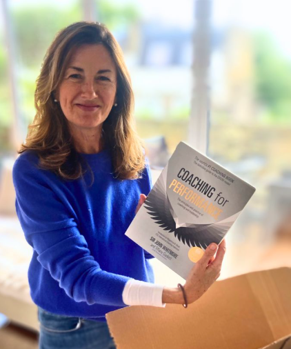 A woman with shoulder-length brown hair, wearing a blue sweater, holding a book titled 'Coaching for Performance,' standing in a bright room with large windows.