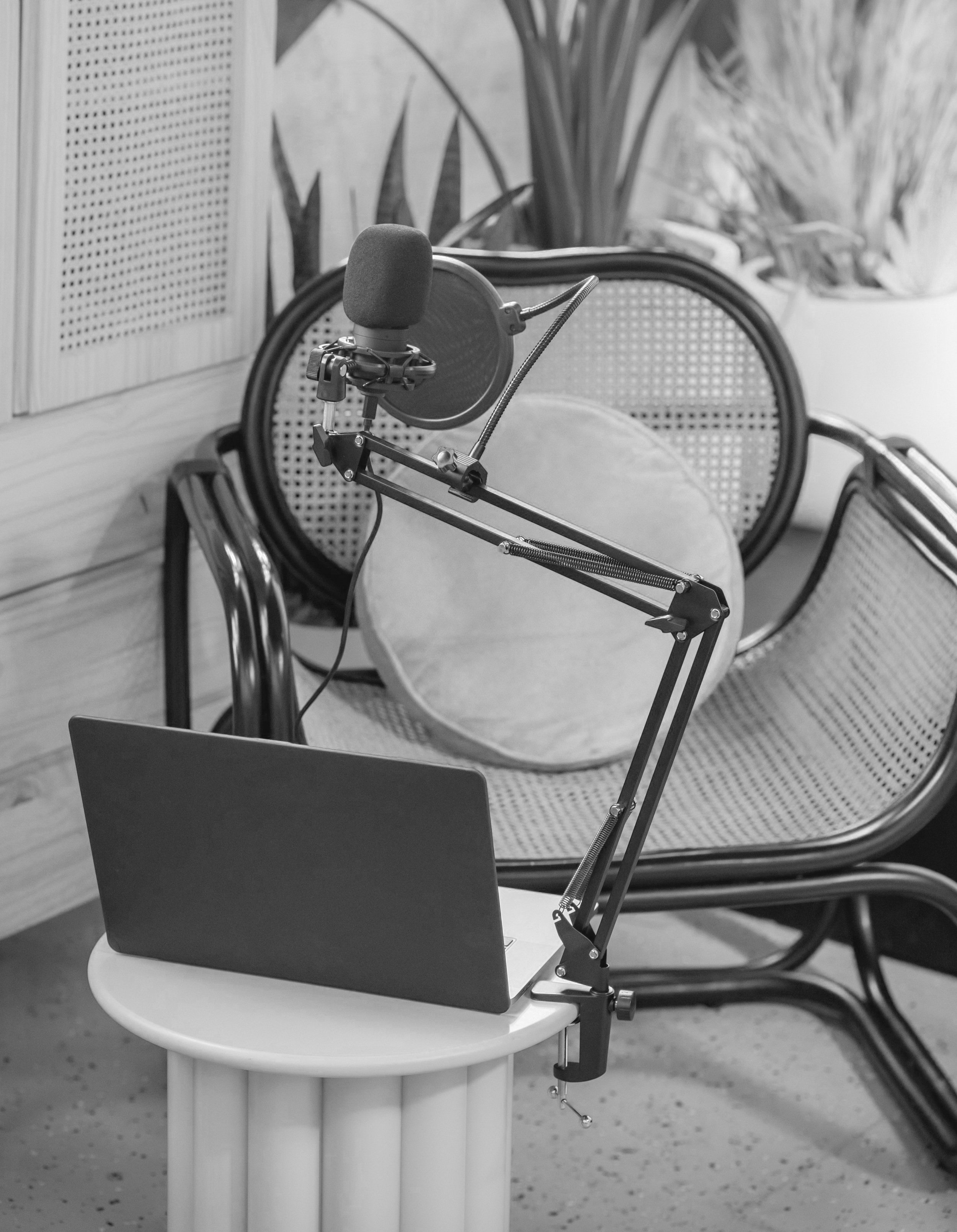 A microphone with a pop filter, a laptop, and a small round table set up for recording or streaming in a room with a wicker chair and potted plants.