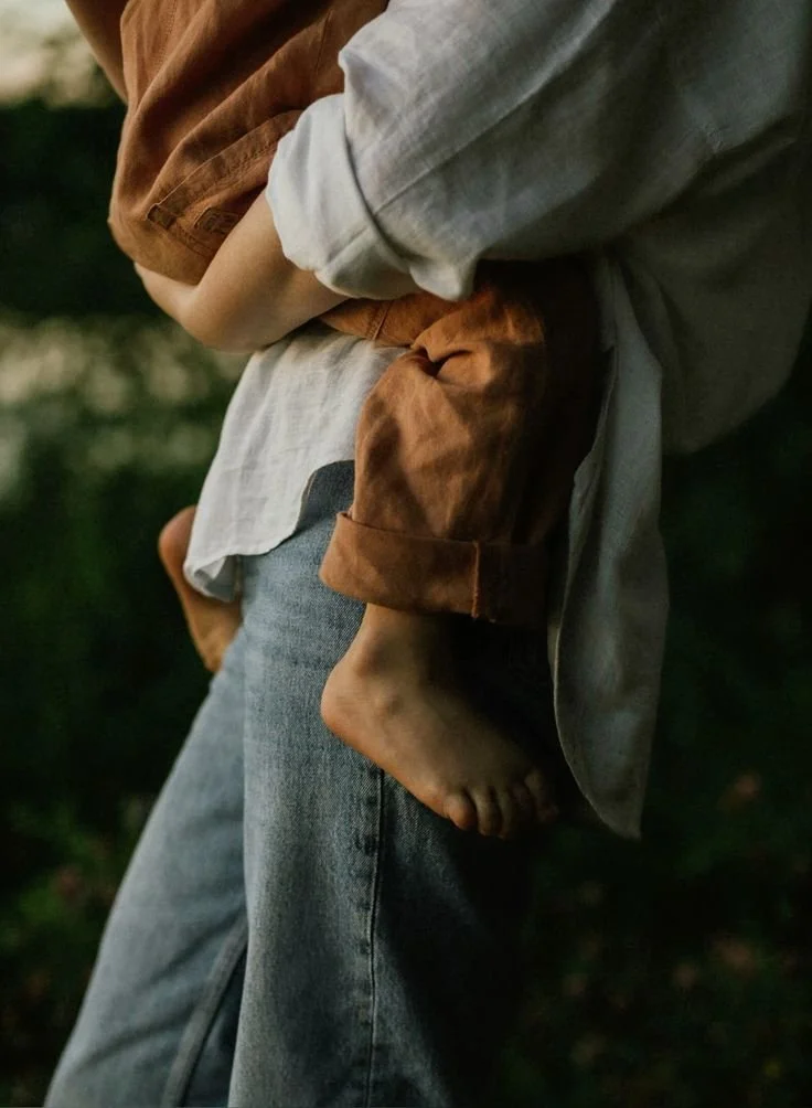 Person carrying a young child outdoors, blurred greenery background, person's jeans and layered shirt visible.