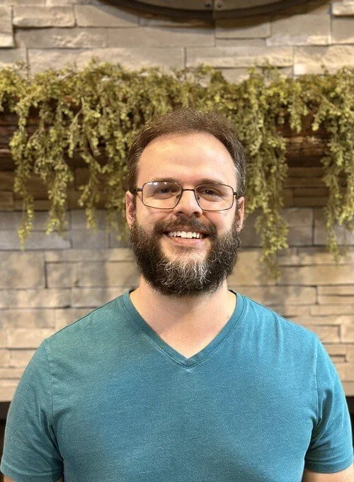 Josh Berg. A man with glasses and a beard smiling, wearing a teal V-neck shirt, standing in front of a brick wall with a wooden shelf and green hanging plants.