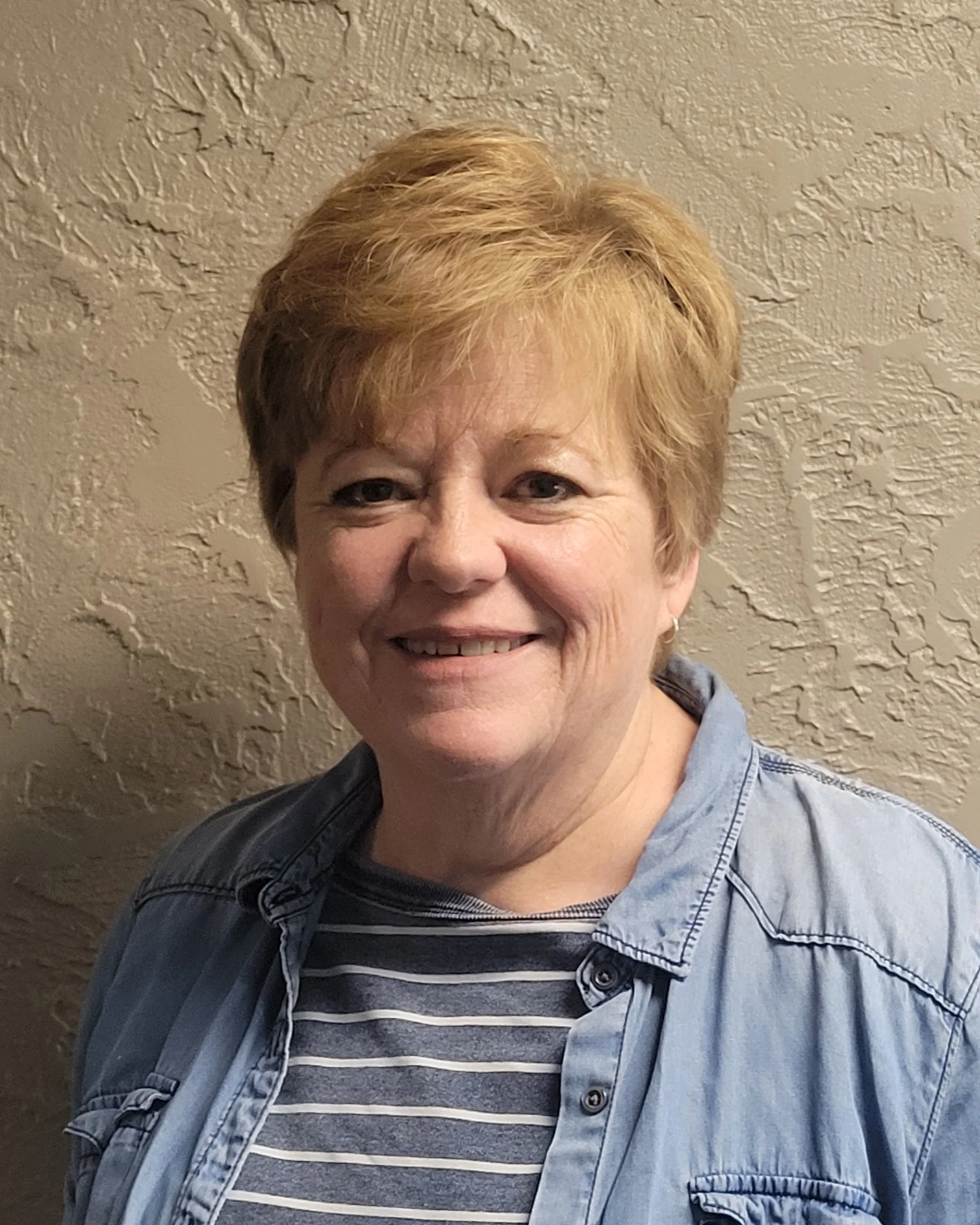 Sherrie Peterson. A smiling woman with short blonde hair, wearing a striped shirt and a denim jacket, standing against a textured beige wall.