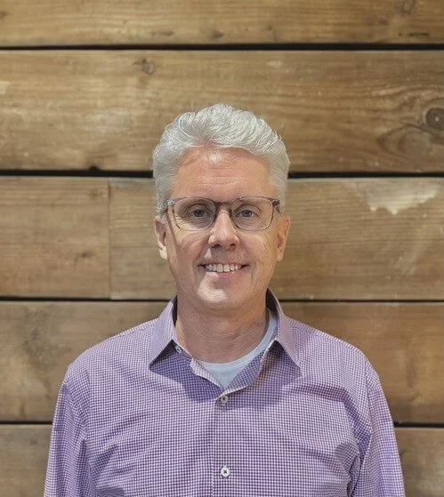 Pastor Kevin Lund. A man with gray hair wearing glasses and a purple checked shirt, smiling in front of a wooden wall.