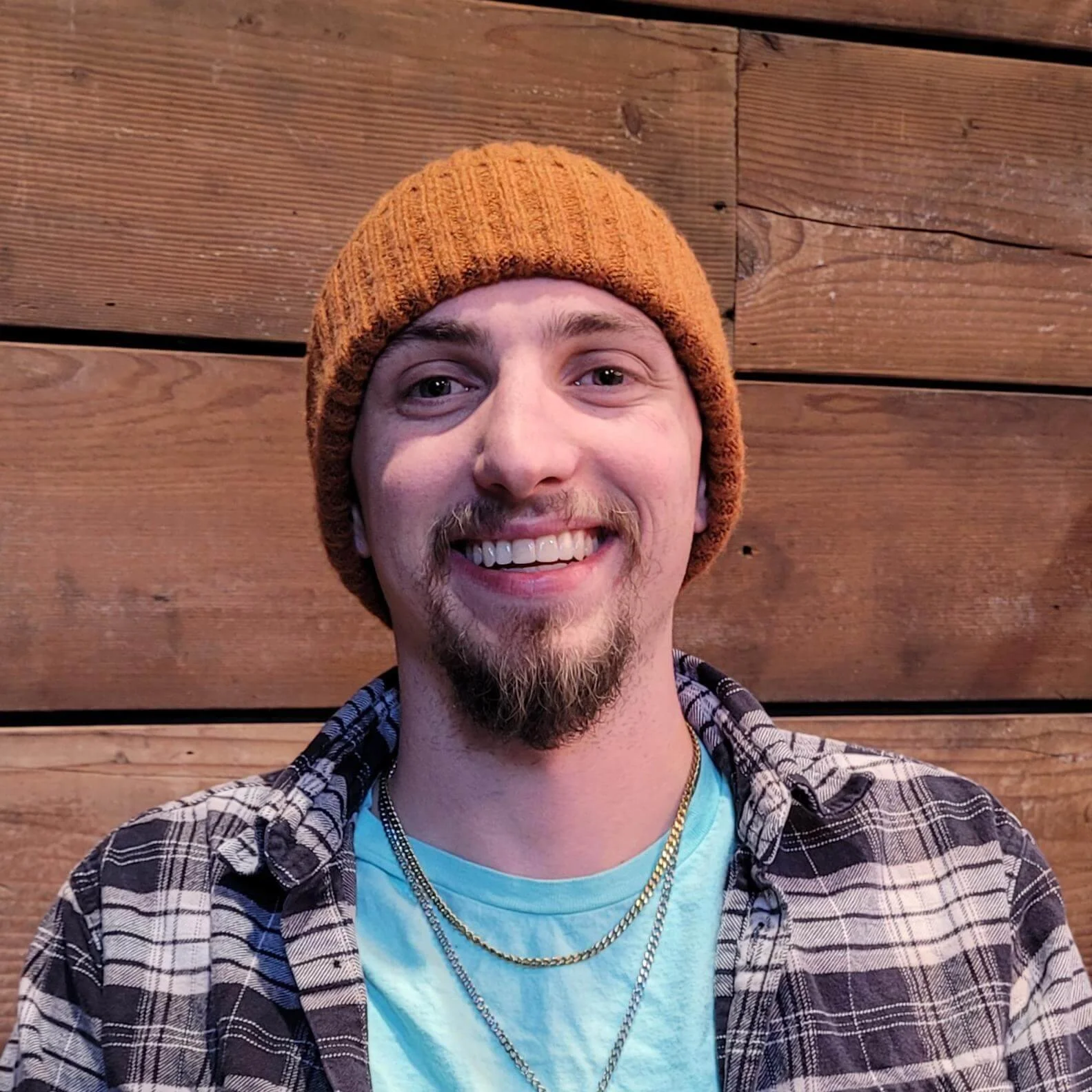 Max Dietz. A smiling young man with a beard, wearing an orange knit beanie, layered necklaces, a mint green t-shirt, and a plaid shirt, standing in front of a wooden wall.