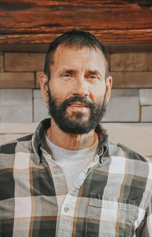 Kevin Erickson. A man with a beard and short dark hair, wearing a plaid shirt over a gray T-shirt, standing indoors with wooden and brick wall background.